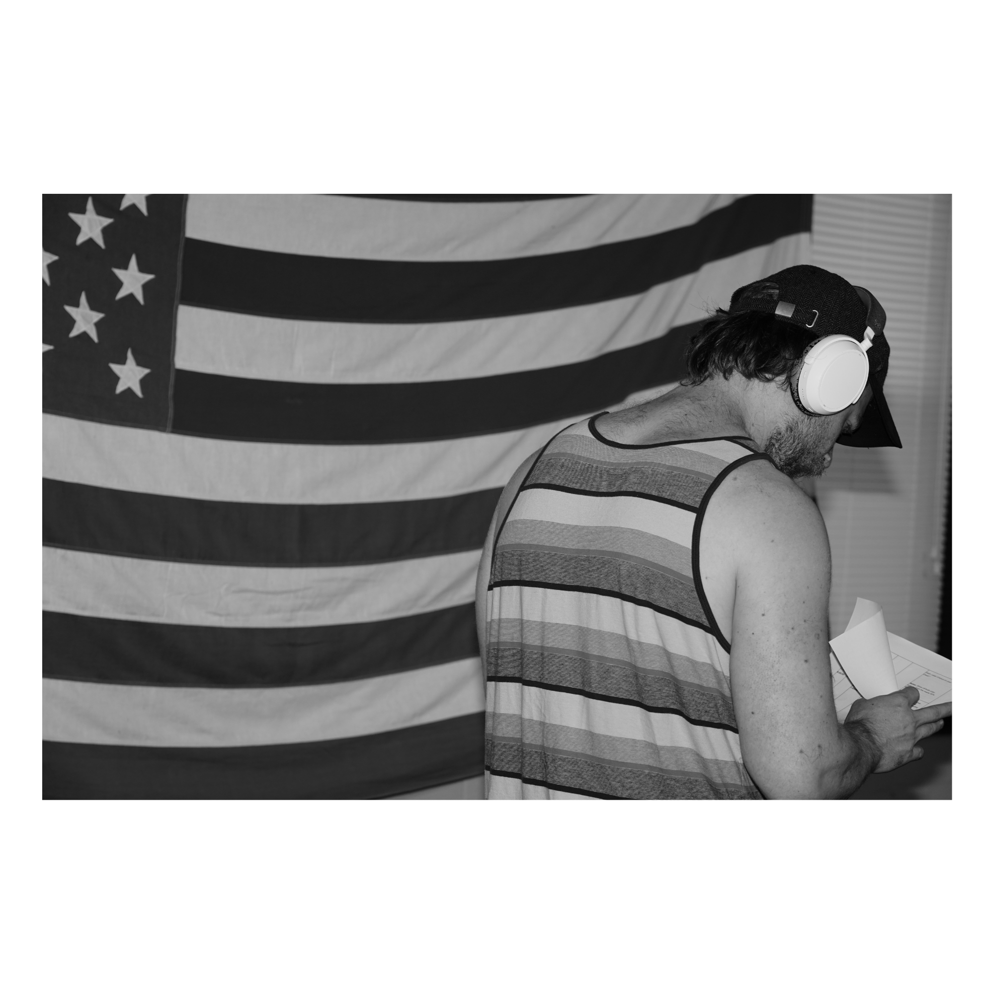 A man wearing striped tank top and headphones holding papers, standing in front of an American flag.