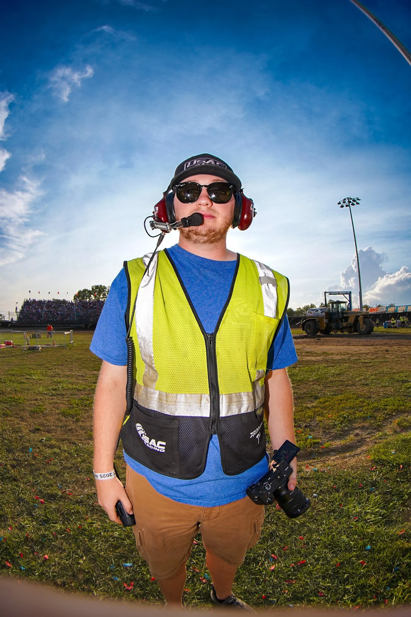 A man standing outdoors at a race event, wearing sunglasses, a black baseball cap, a yellow safety vest, holding a camera, with a crowd and fleet of construction vehicles in the background.