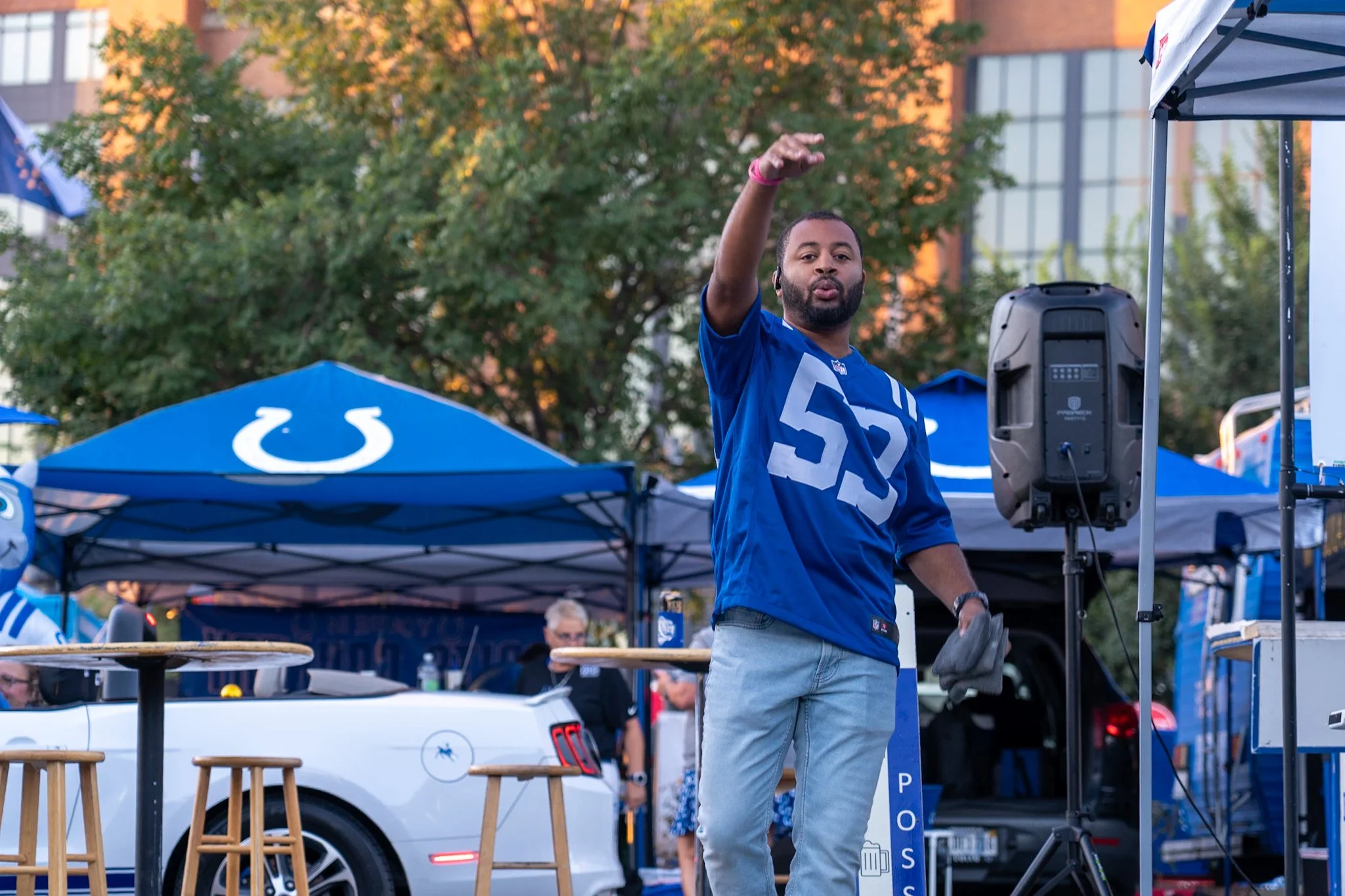 A man in an Indianapolis Colts football jersey waving at an outdoor event with tents and people in the background.