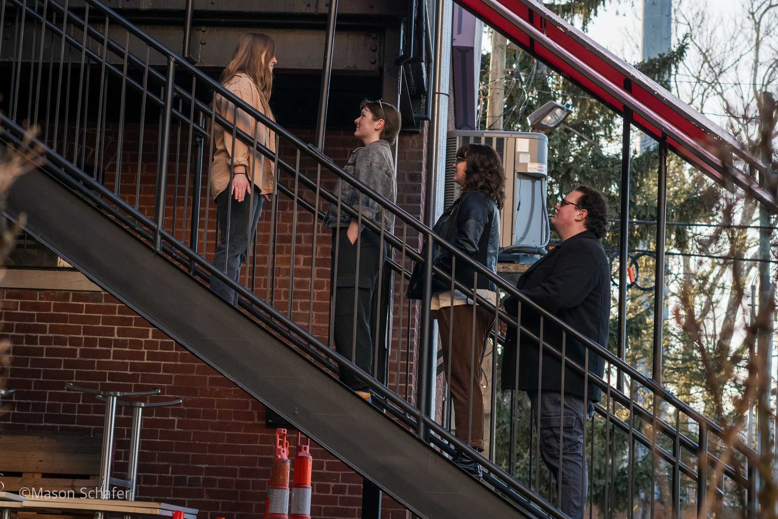 Four people stand on an outdoor staircase, two women and two men, engaging in conversation. One woman is on the upper level, while the other three are on the lower level, including the man in black. The staircase is metal with vertical railings, agai