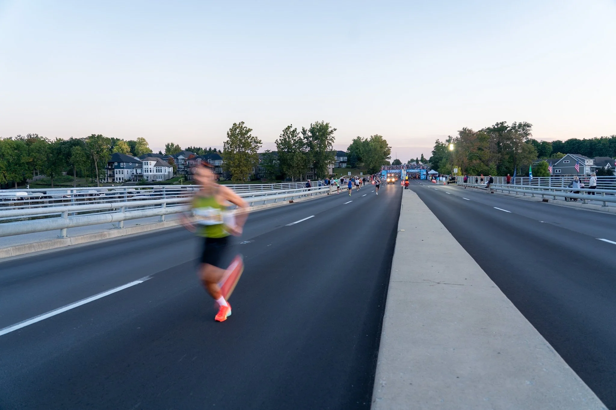 Blurred male runner participating in a race on a paved road, with houses and trees in the background, and tents and a finish line in the distance.