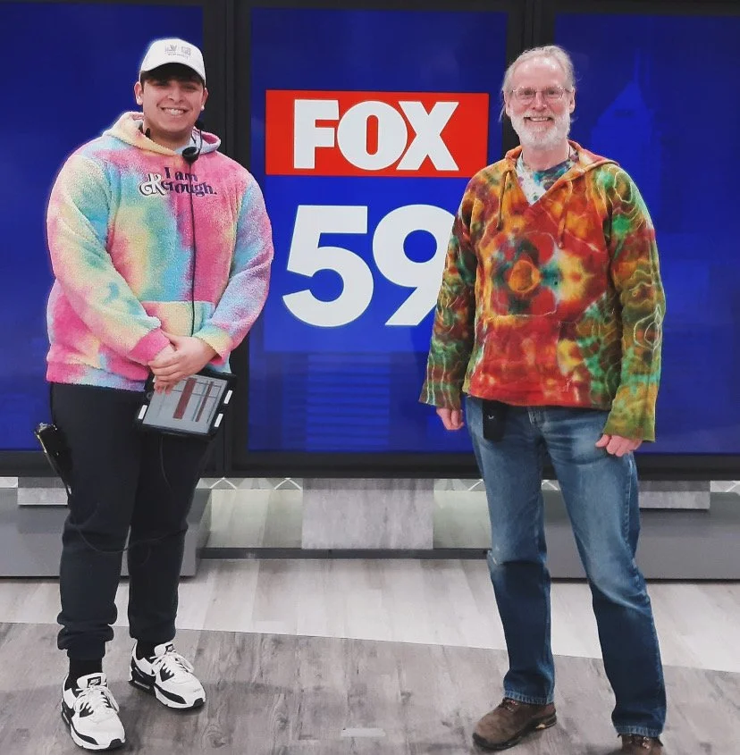 Two men stand in front of a large FOX 59 news screen; one wears a colorful tie-dye hoodie, black pants, and sneakers, while the other wears a multicolored tie-dye jacket, jeans, and boots.