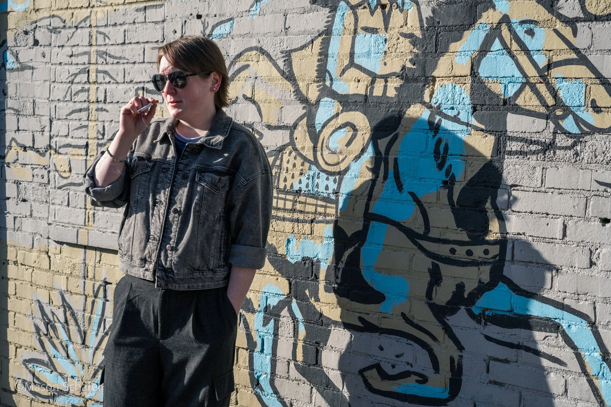 A young woman with short brown hair, wearing black sunglasses, a denim jacket, and dark pants, standing beside a brick wall with colorful graffiti art. The graffiti features bold black outlines and shades of blue and beige, depicting a stylized face 
