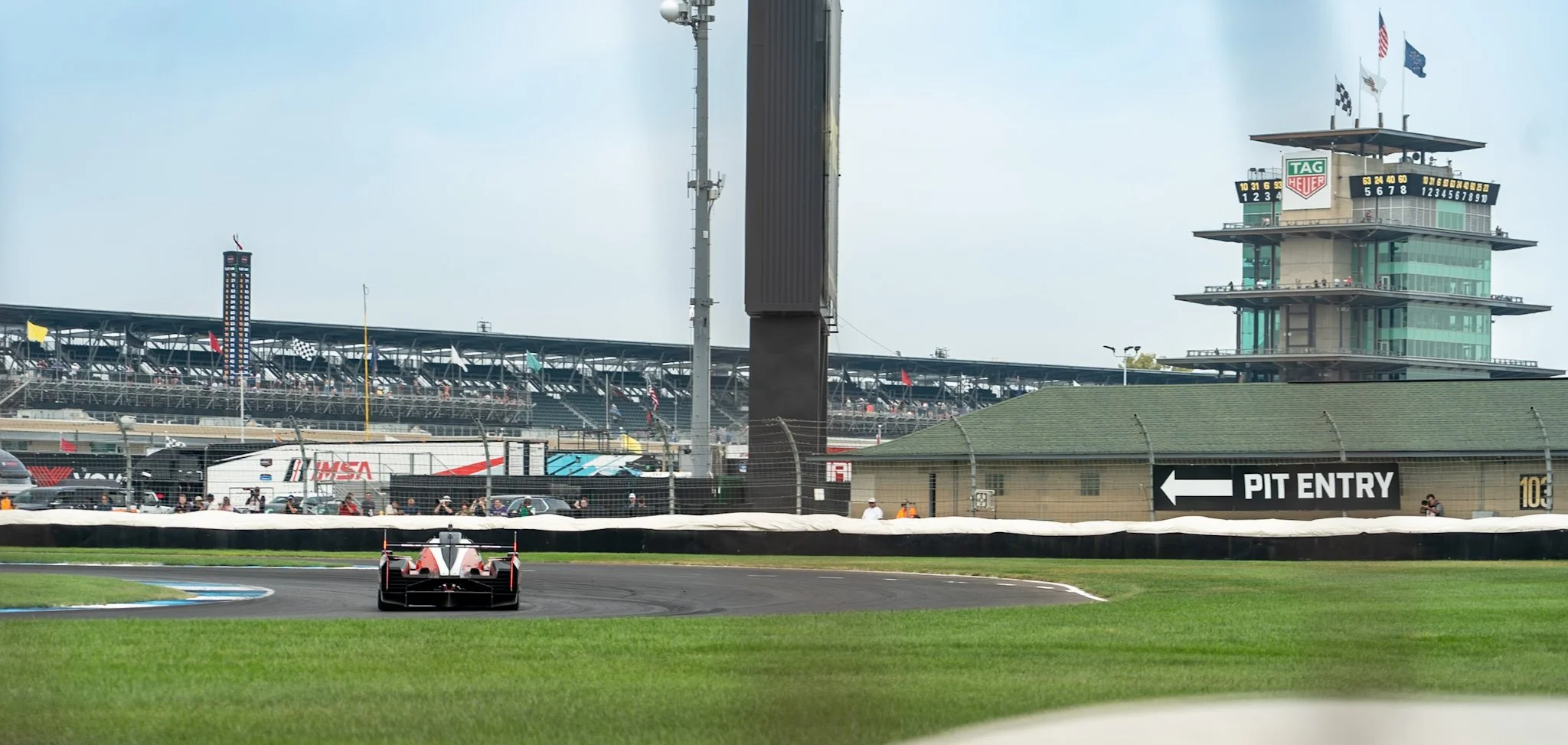 A racing car on a race track at a motorsports stadium, with the grandstands and control tower in the background, and a sign pointing to the pit entry on the right.