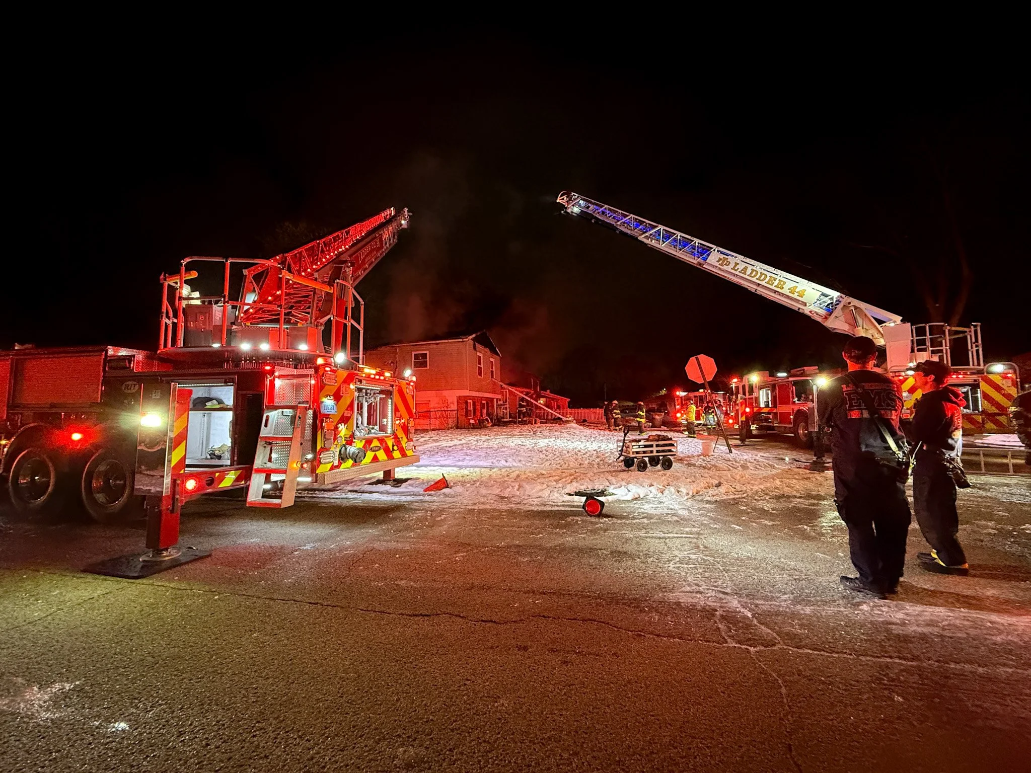Nighttime scene with firefighters and two fire trucks, one with an extended ladder, in a residential neighborhood. Firefighters are standing on the street, and the area is illuminated by emergency lights with some snow on the ground.