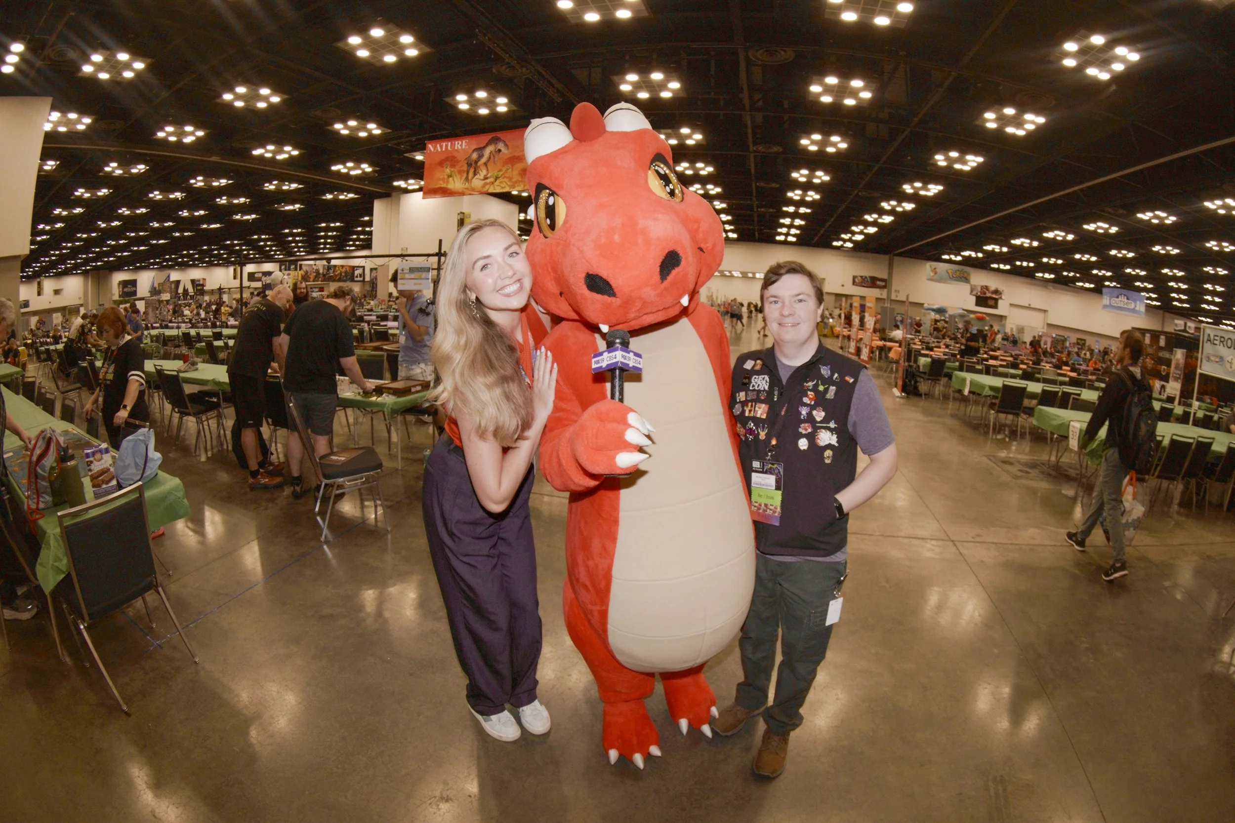 Two people dressed as a woman and a young man standing with a person in a red dinosaur mascot costume at a convention or expo in a large hall filled with tables and attendees.