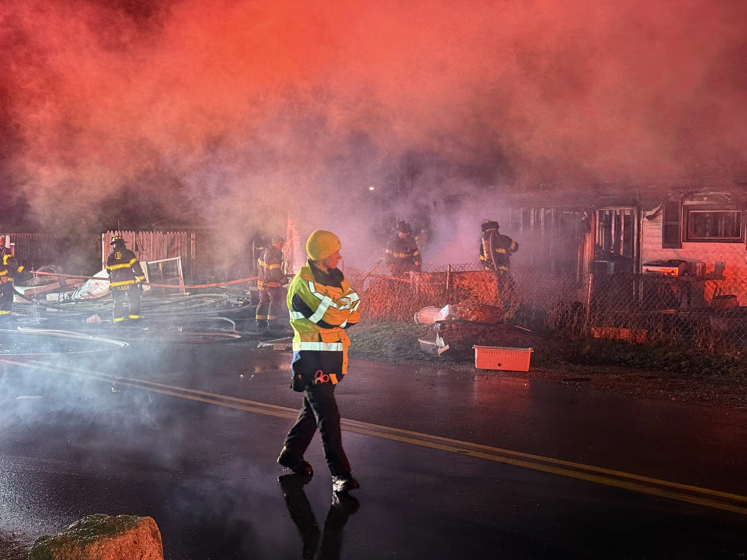 Firefighters working to extinguish a house fire at night, with smoke and flames visible, and one firefighter standing in the street.