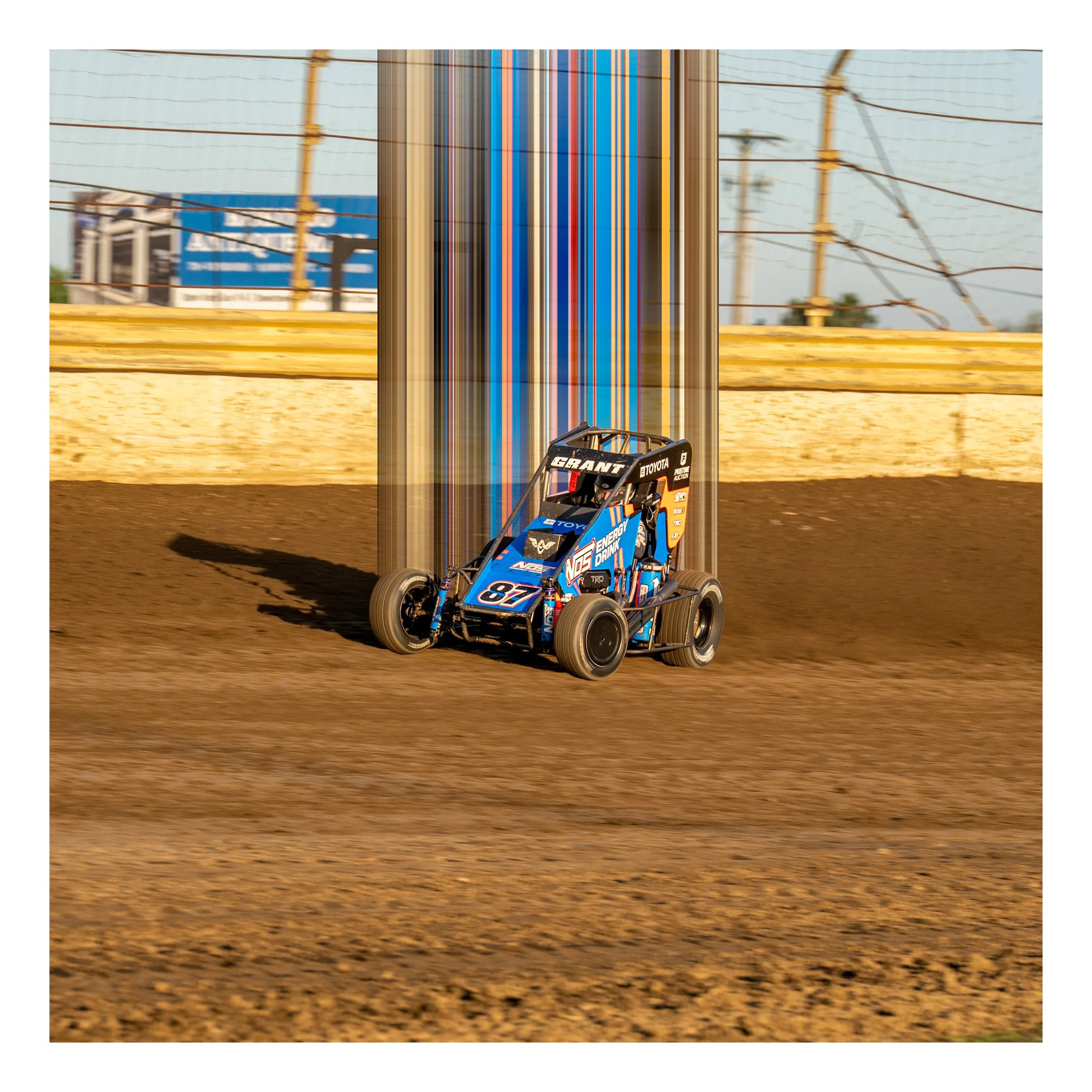 Close-up of a sprint car on a dirt race track, with a blurred background of a billboard and power lines.