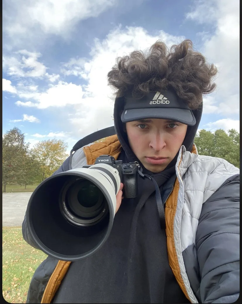 Young person outdoors taking a selfie, pointing a camera towards the lens, wearing a black Adidas visor, a hooded jacket with a brown front panel, and a black hoodie, with trees and a partly cloudy sky in the background.