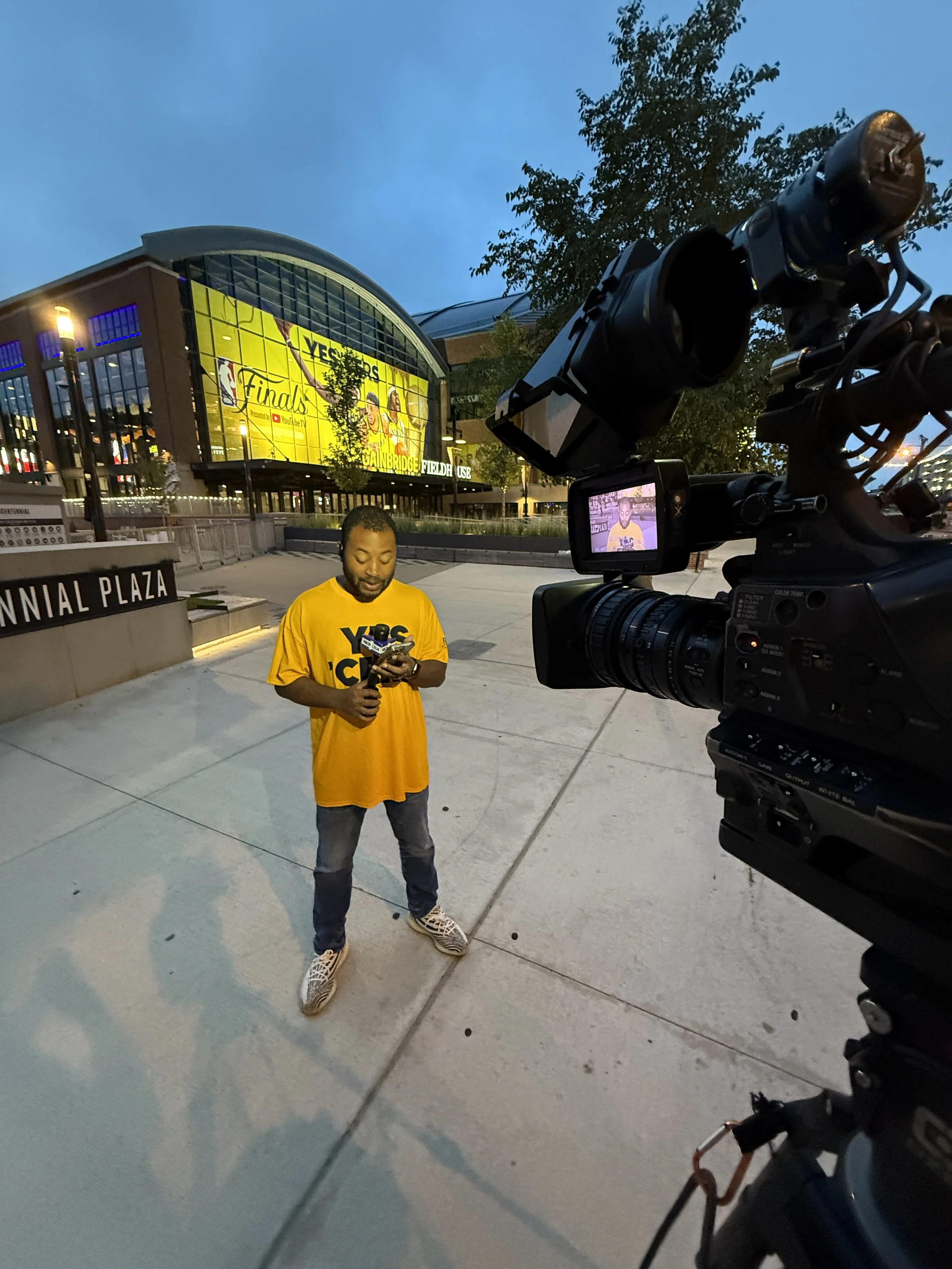 A man in a yellow shirt with 'YBC Champs' holding a phone, being filmed by a video camera outside in front of a stadium labeled 'Fennial Plaza' during early evening.