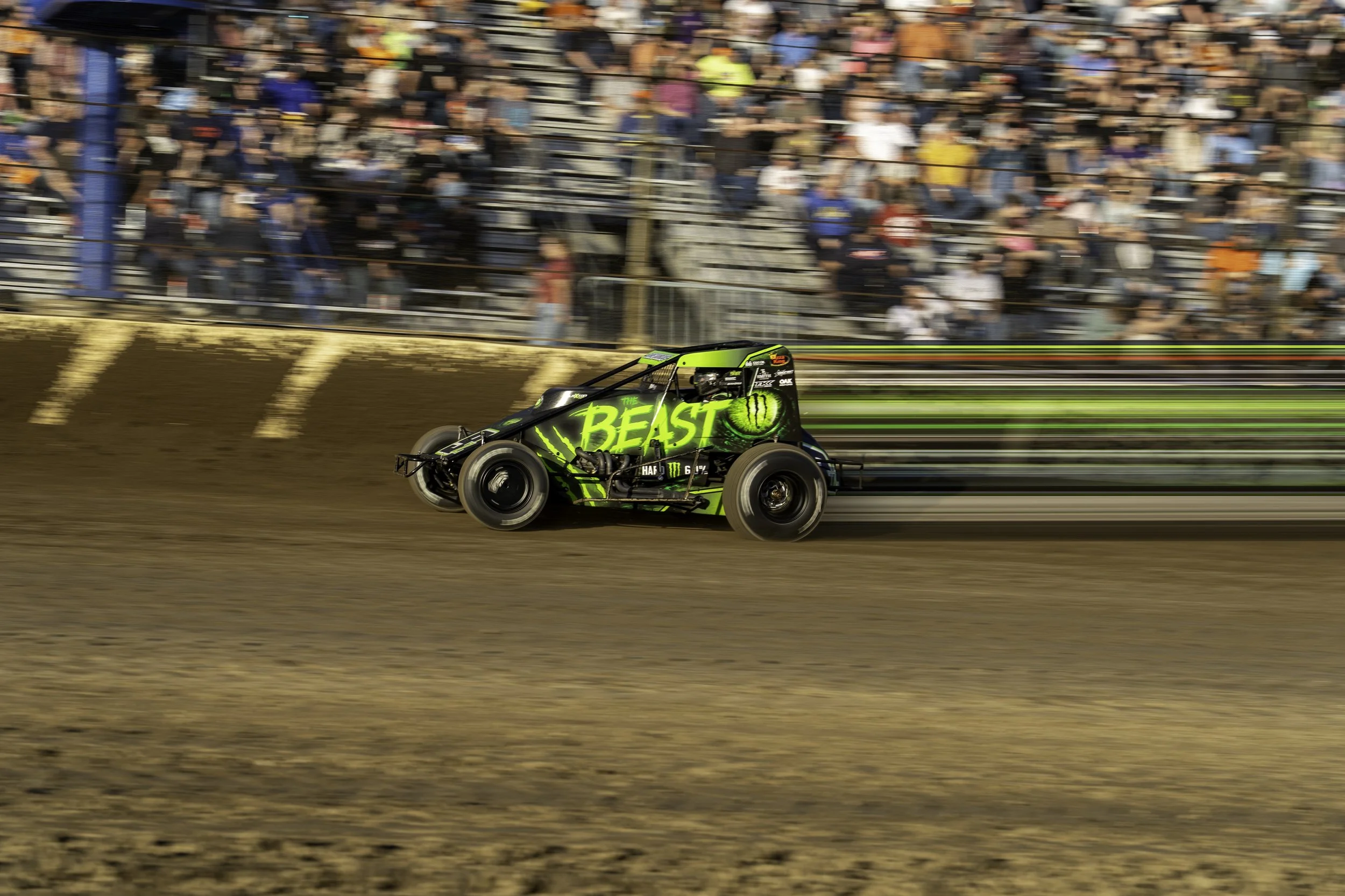 A dirt track racecar with green and black colors, featuring the words 'The Beast' and a large green eye design, moving at high speed with a blurred audience in the background.
