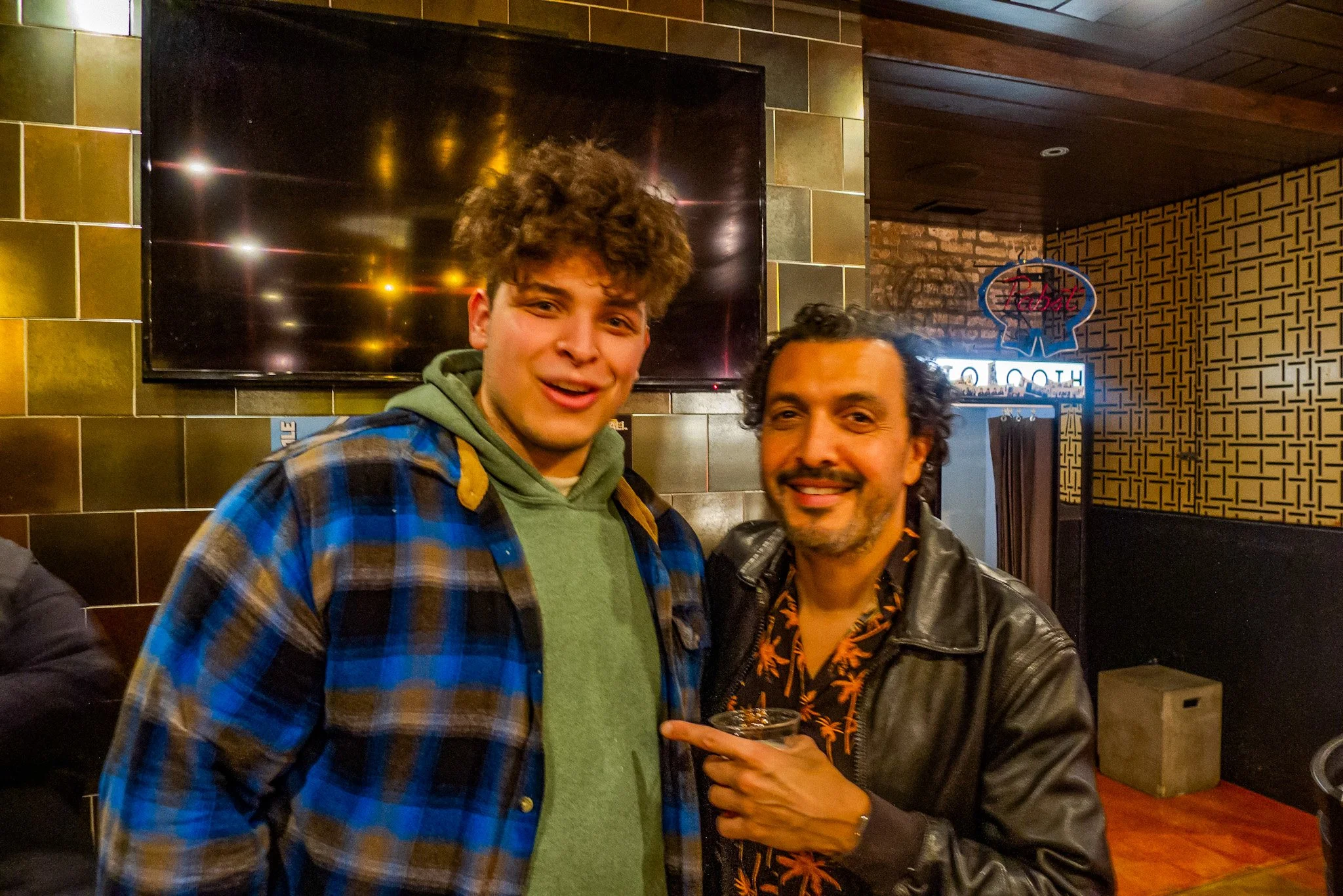 Two men smiling and posing for a photo inside a restaurant or bar, with a TV screen and decorative wall in the background.