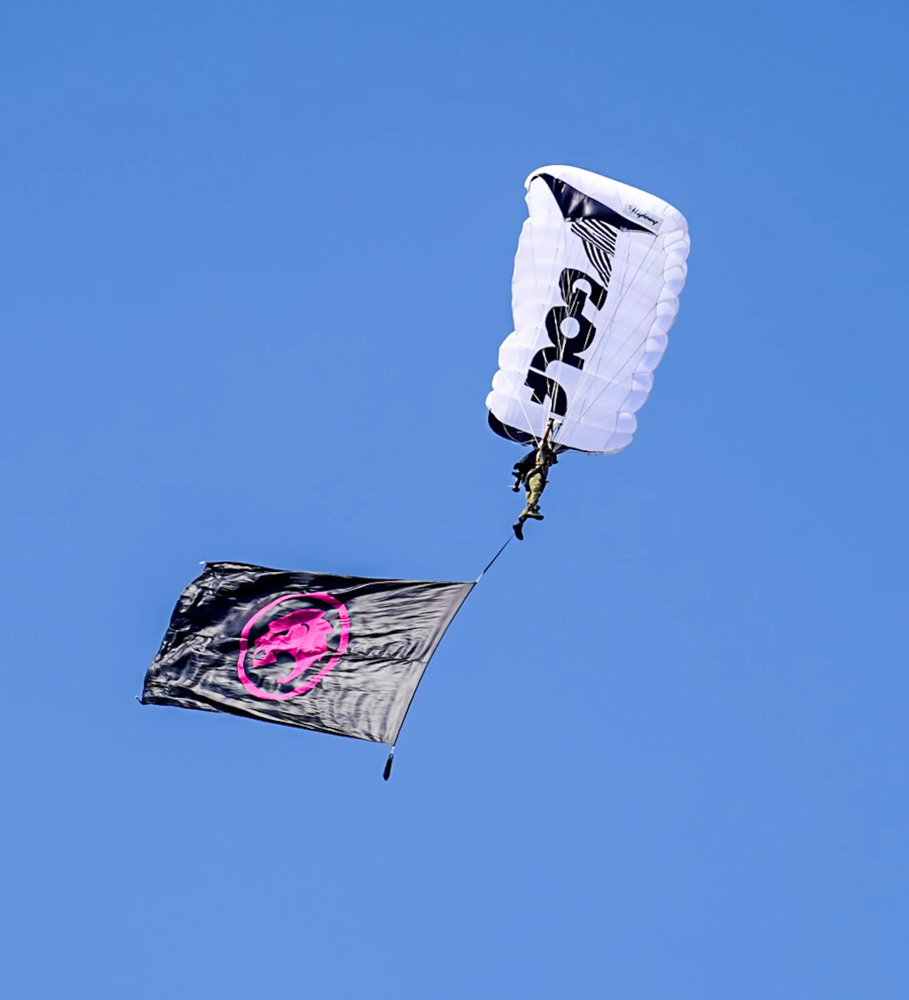 A person paragliding with a white parachute and a black flag with a pink skull symbol underneath.