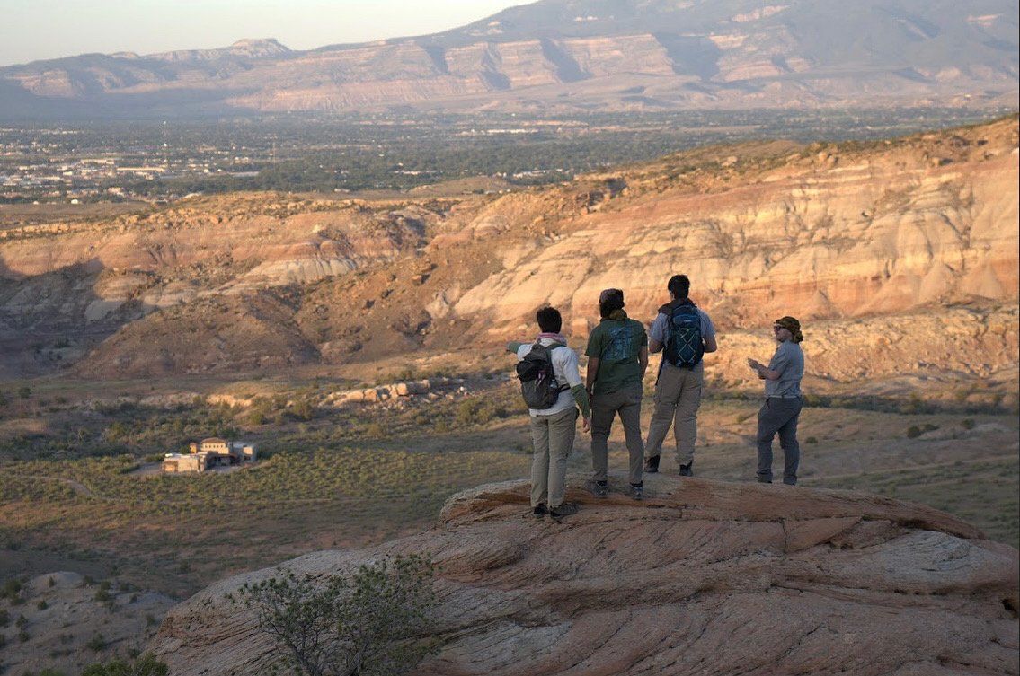 Four people standing on a rocky ledge overlooking a desert canyon with mountains in the distance, some holding maps or papers.