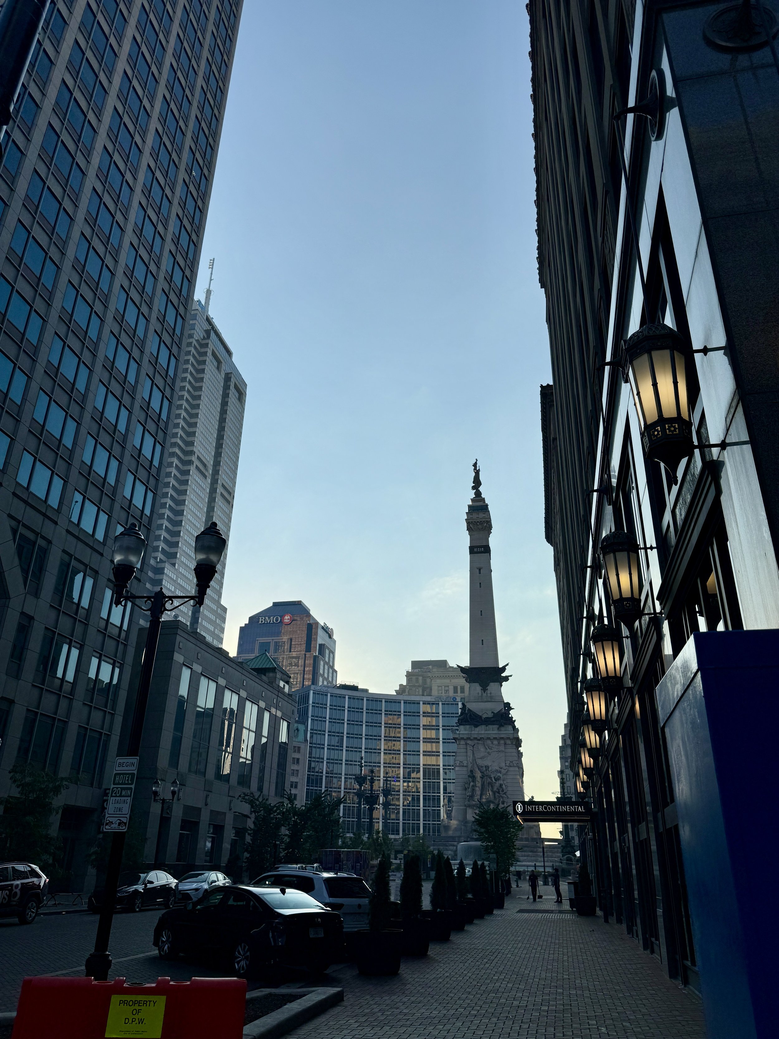 City street with tall buildings, parked cars, and a monument with a statue on top, under a clear blue sky.
