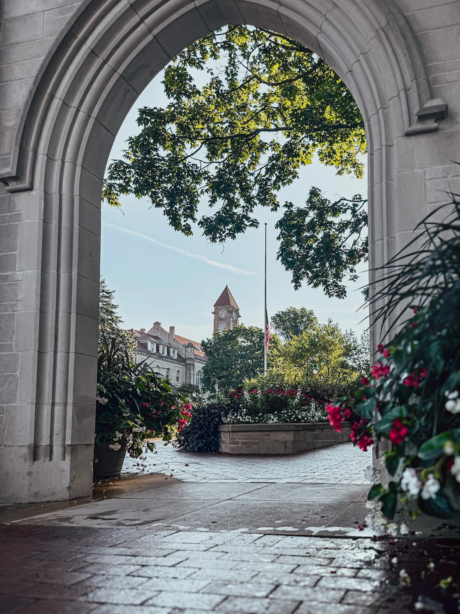 View through a stone arch into a landscaped courtyard with a clock tower in the background, surrounded by trees and flowering bushes.