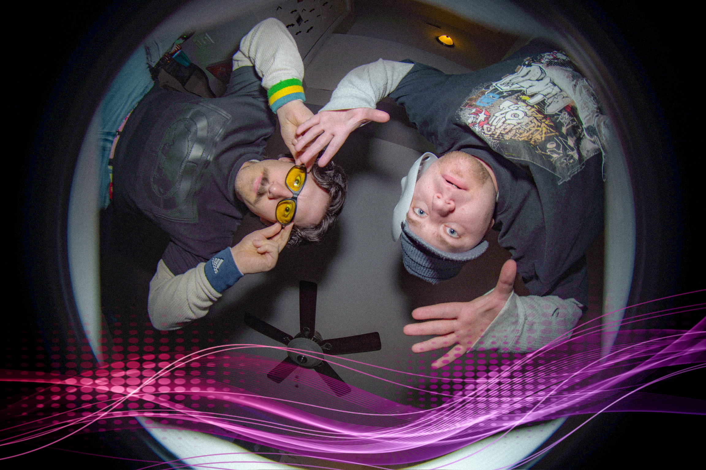 Two young men lying on the floor and looking up at the camera through a fisheye lens, with a pink digital wave pattern overlay at the bottom of the image.