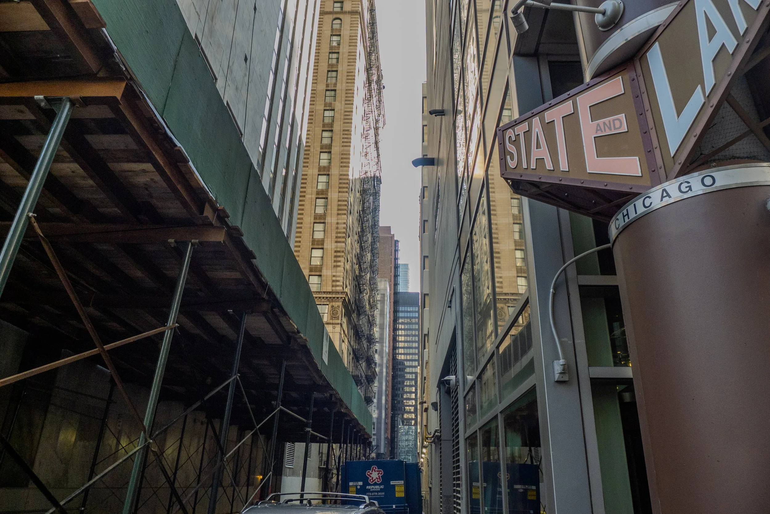 View of a narrow city alleyway with tall buildings, construction scaffolding, and a vintage sign that says 'STATE' and 'CHICAGO' on the sidewalk.