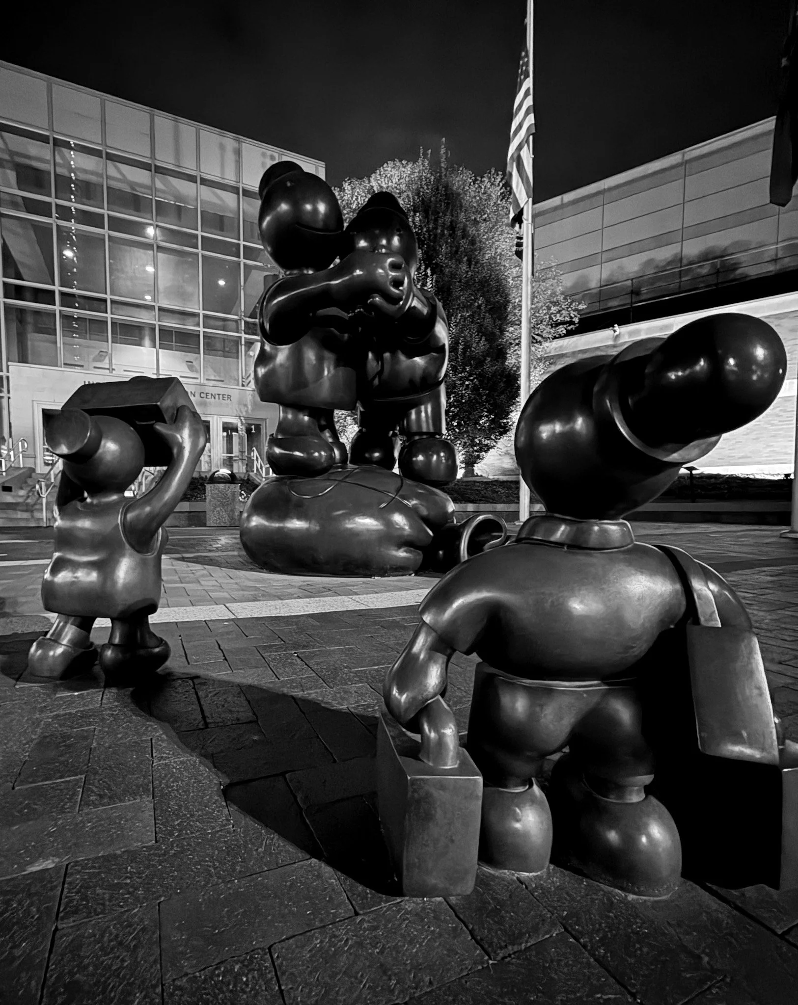 Nighttime black and white photo of a public art installation featuring sculptures of children holding lunchboxes, with one removed and placed on the ground, and a large abstract figure in the background. An American flag is visible in the background,