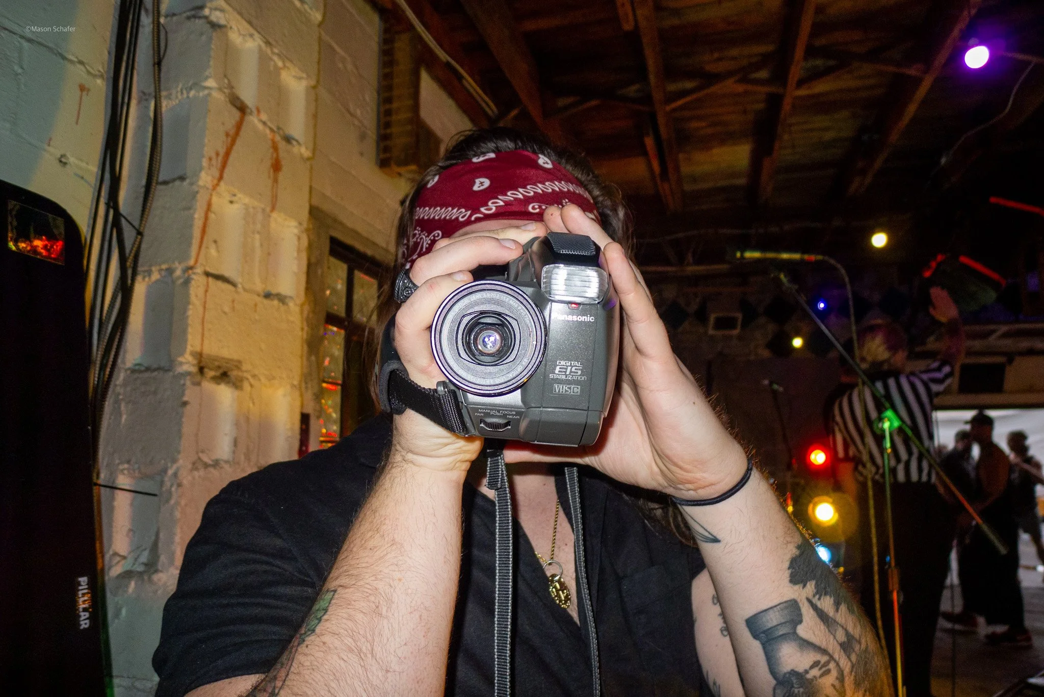 Person holding a camera pointed at the viewer, with a bandana on their head, tattoos on their arms, and a necklace with a pendant, at a lively indoor event with stage lights and people in the background.