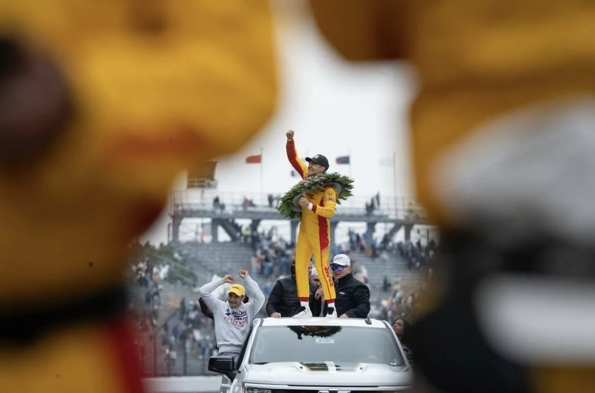 Race car driver celebrating on top of a vehicle, holding a wreath, with people cheering in the background, framed by blurred yellow and black objects in the foreground.