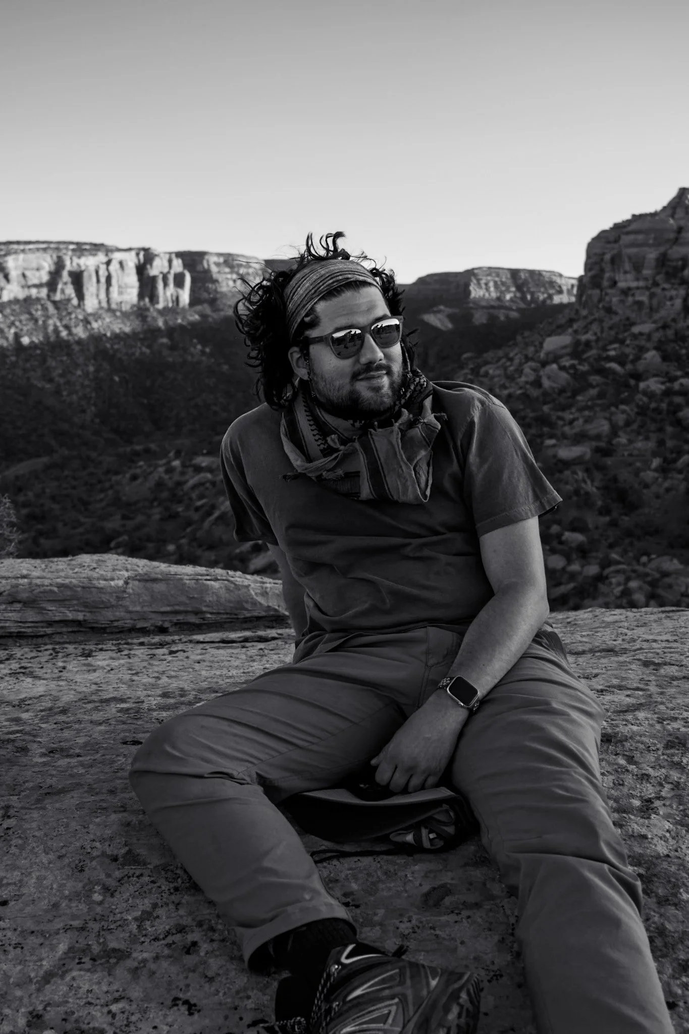 A man sitting on a rock ledge in a desert landscape with cliffs in the background, wearing sunglasses, a cap, a scarf, a watch, and outdoor clothing, captured in black and white.