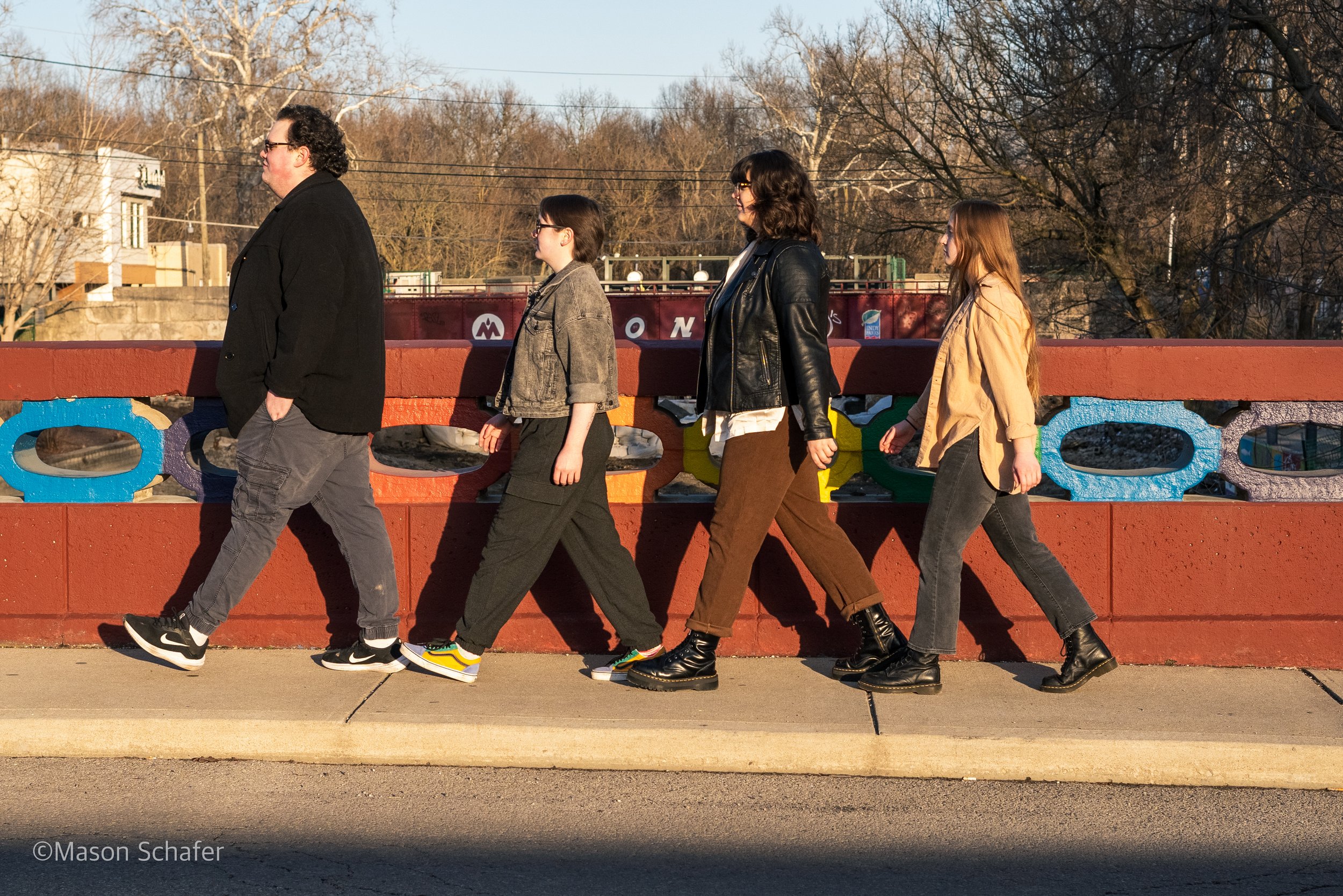 Five people walking in a line across a sidewalk, still in a military style formation, on a sunny day, with a colorful painted bridge and leafless trees in the background.