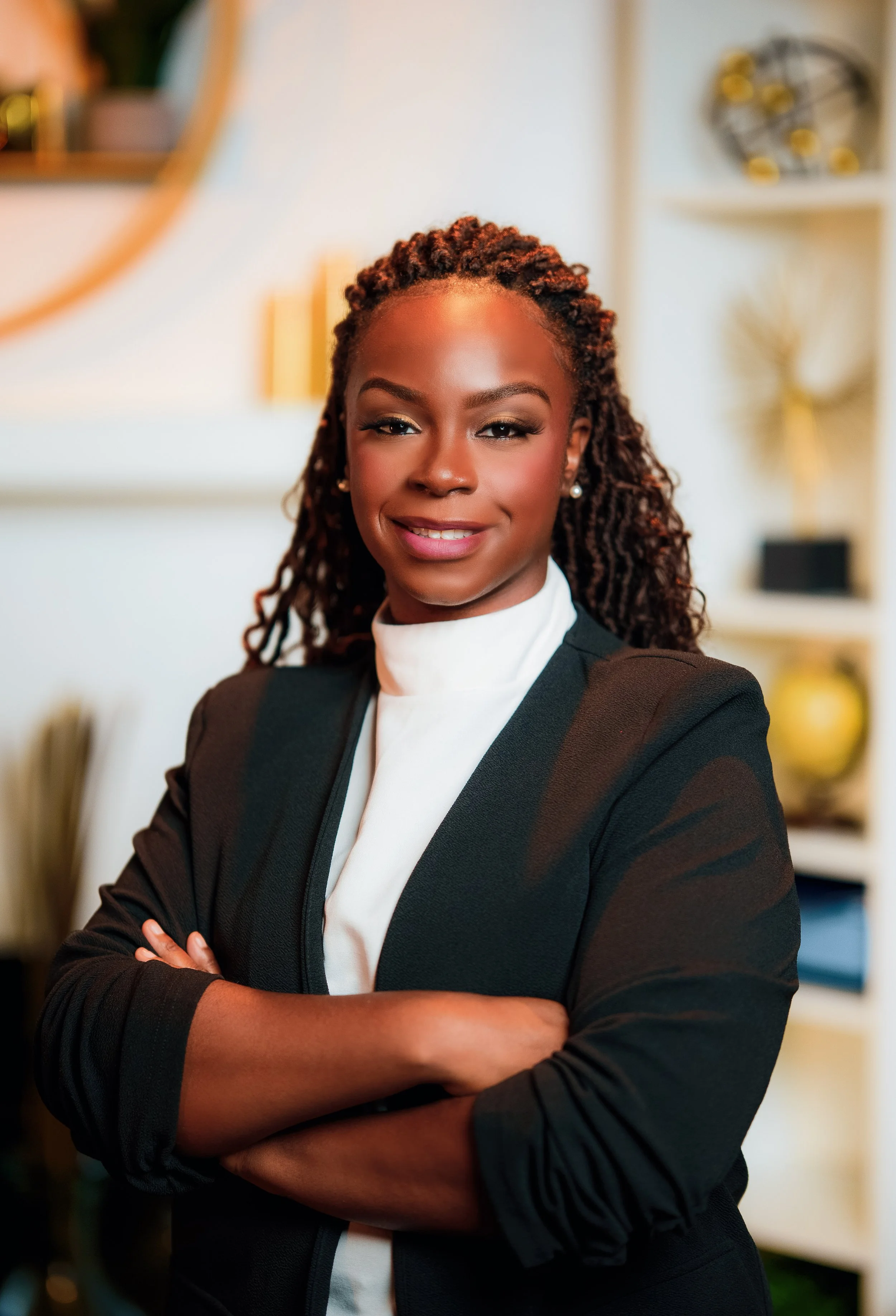 A confident woman with dark hair styled in twists, wearing a black blazer over a white blouse, standing with arms crossed in a modern, stylish interior.