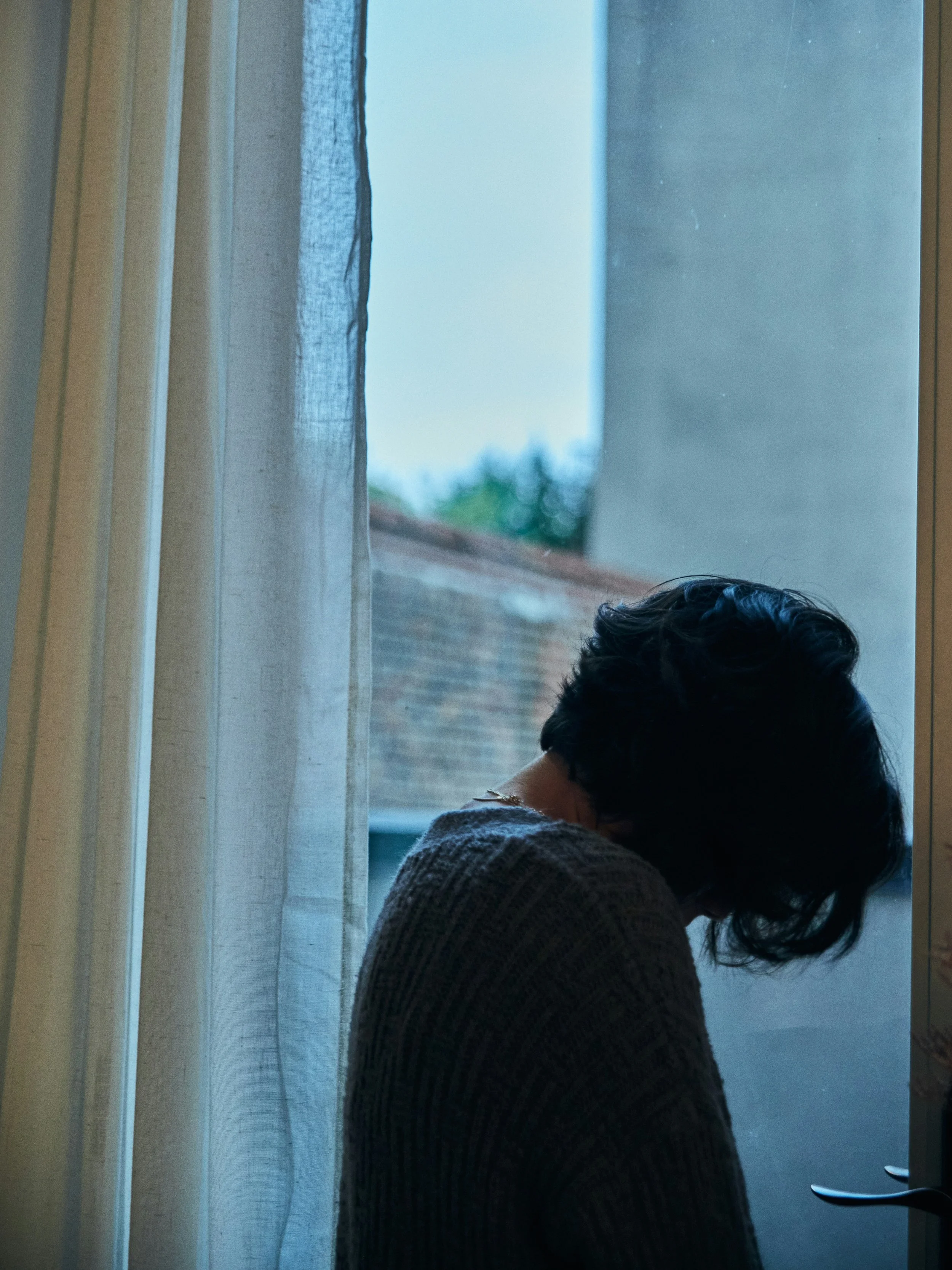 A person with dark hair, wearing a knitted sweater, sitting by a window with sheer curtains, looking downward.