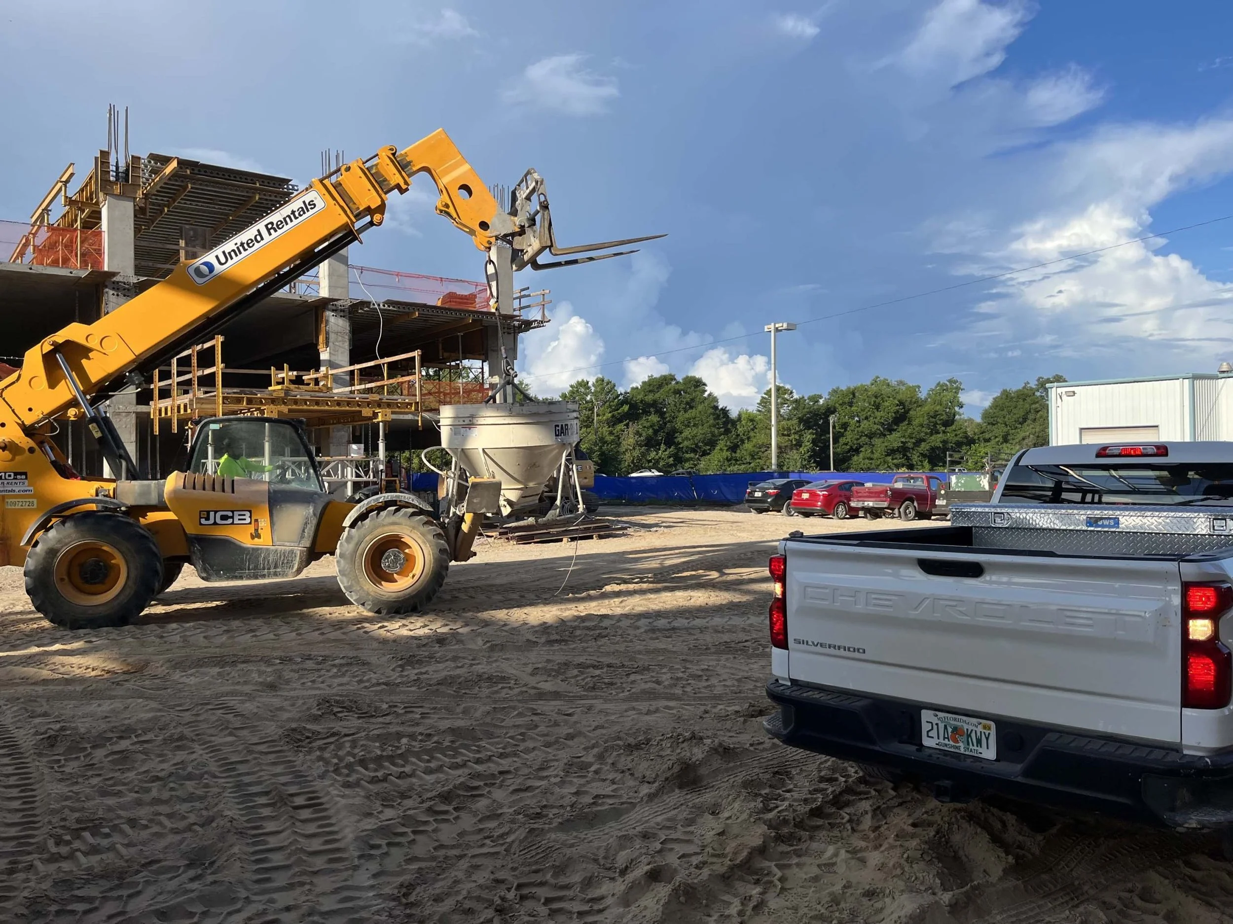 Construction site with a yellow forklift lifting a bucket, partially built structure in the background, parked vehicles including a white Silverado truck in the foreground, and a partly cloudy blue sky.