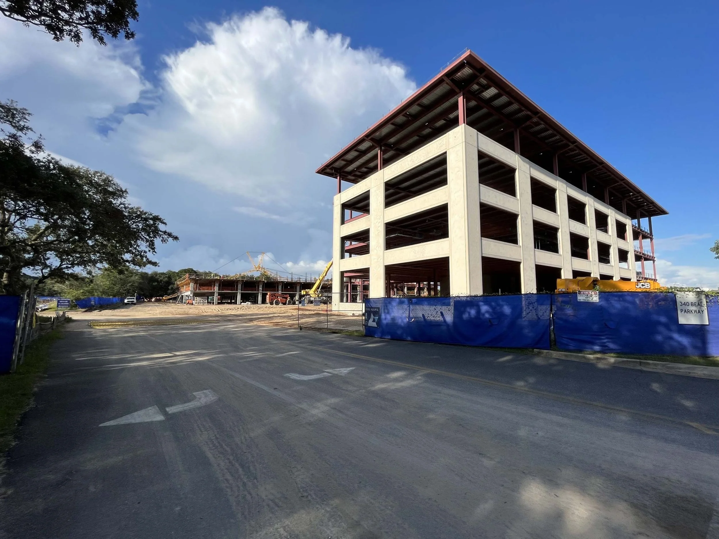 Construction site with a partially built multi-story concrete building, construction equipment, and blue safety fencing, under a partly cloudy sky.