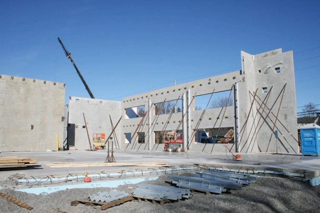 Construction site with partially built concrete walls, supporting poles, a blue portable toilet, and a crane under a clear blue sky.