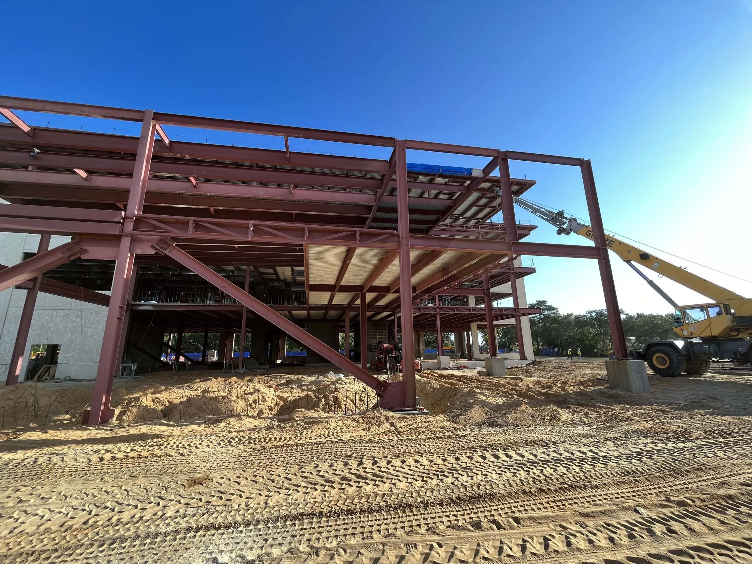 Construction site with a steel building frame under construction, yellow crane lifting materials, and dirt ground with tire tracks.
