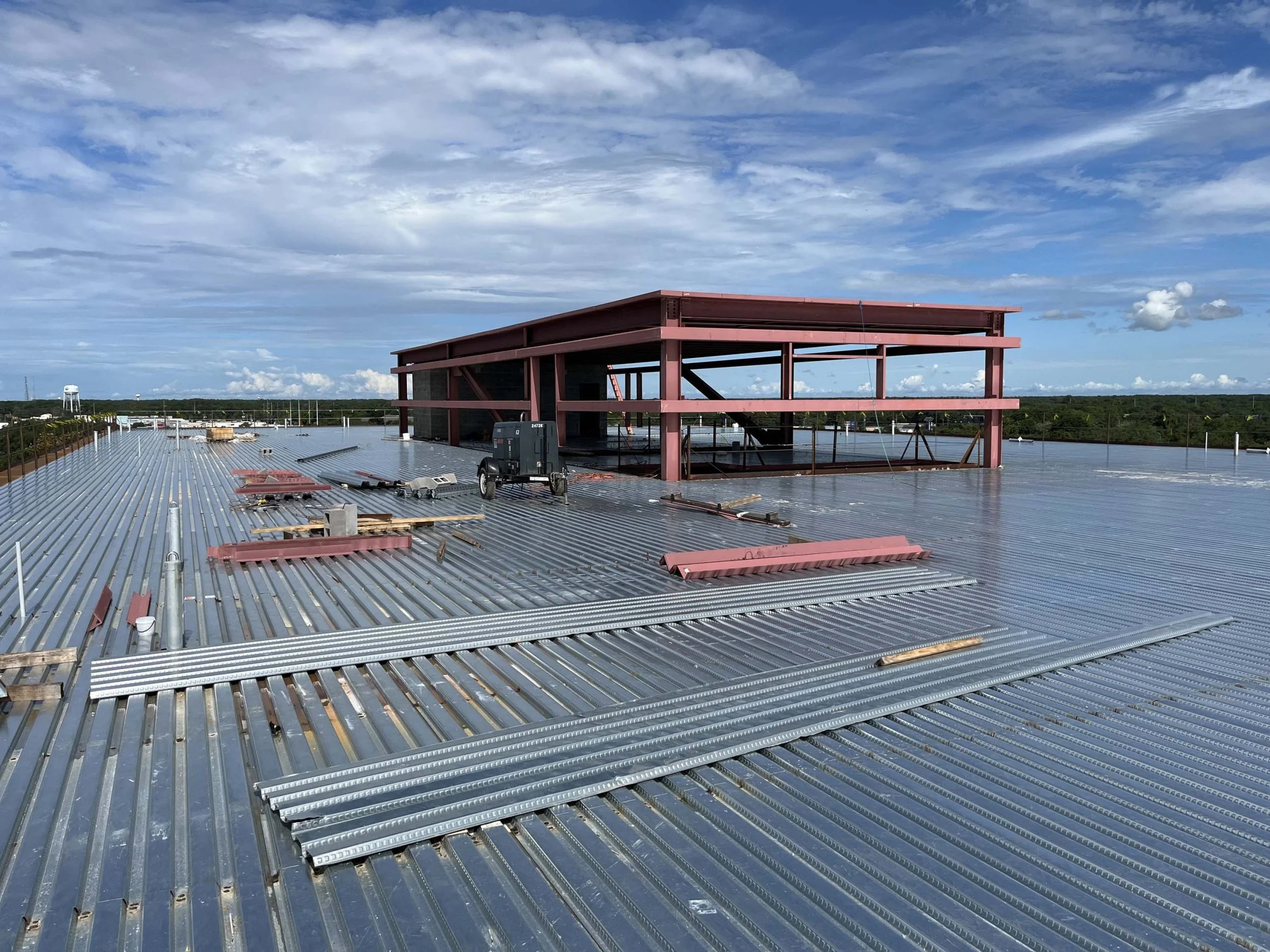 Construction site on a large metal roof with a partially built steel framework structure in the background under a partly cloudy sky.