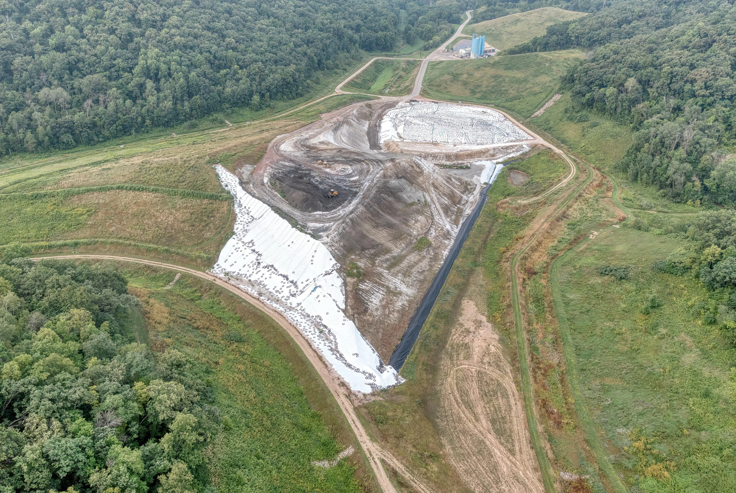 Aerial view of a construction site in a green, forested area with dirt roads and machinery working on earth, with a blue silo structure in the background.