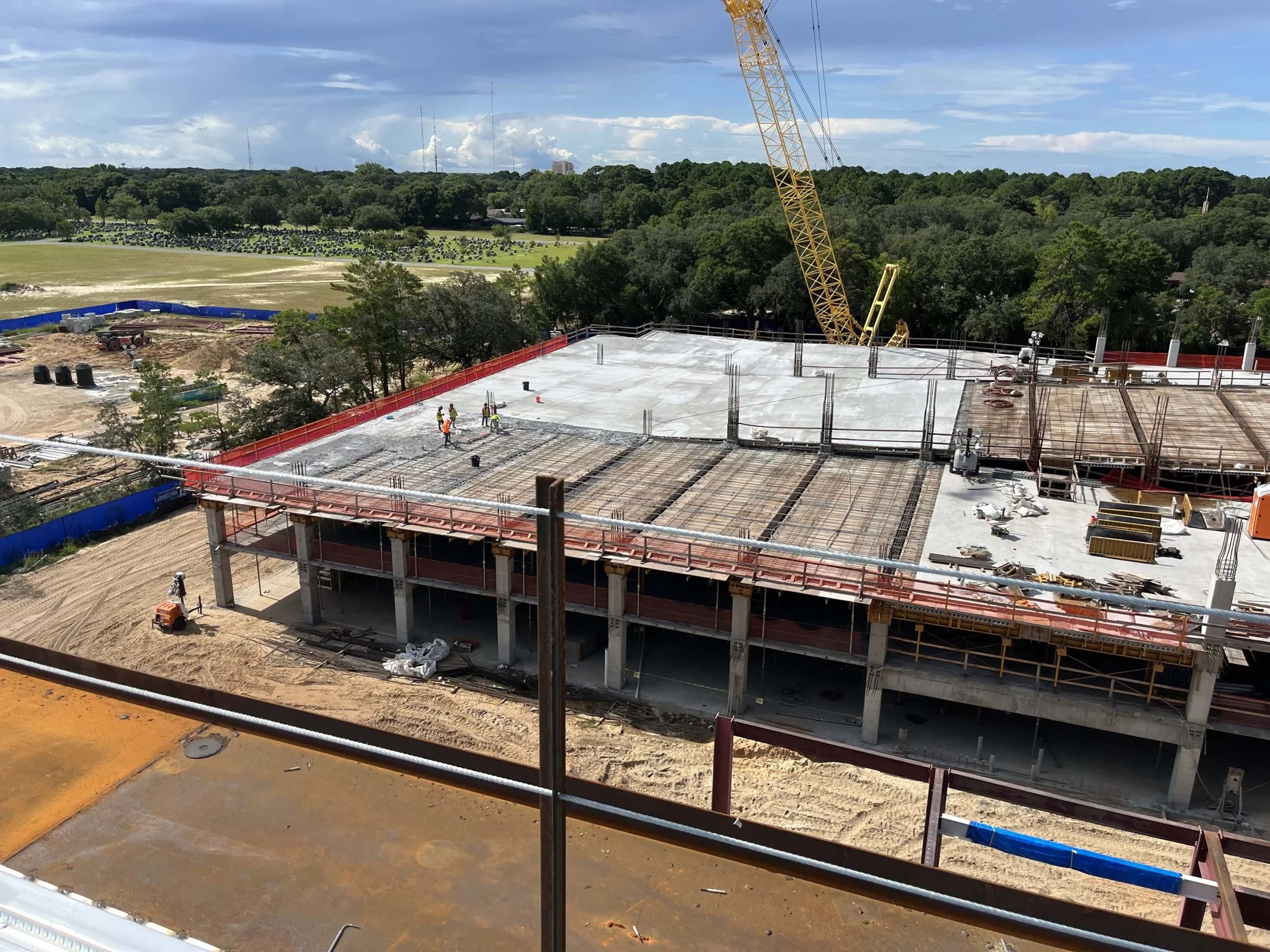 Construction site with workers laying concrete on a building's top floor, a yellow crane in the background, and trees in the distance.