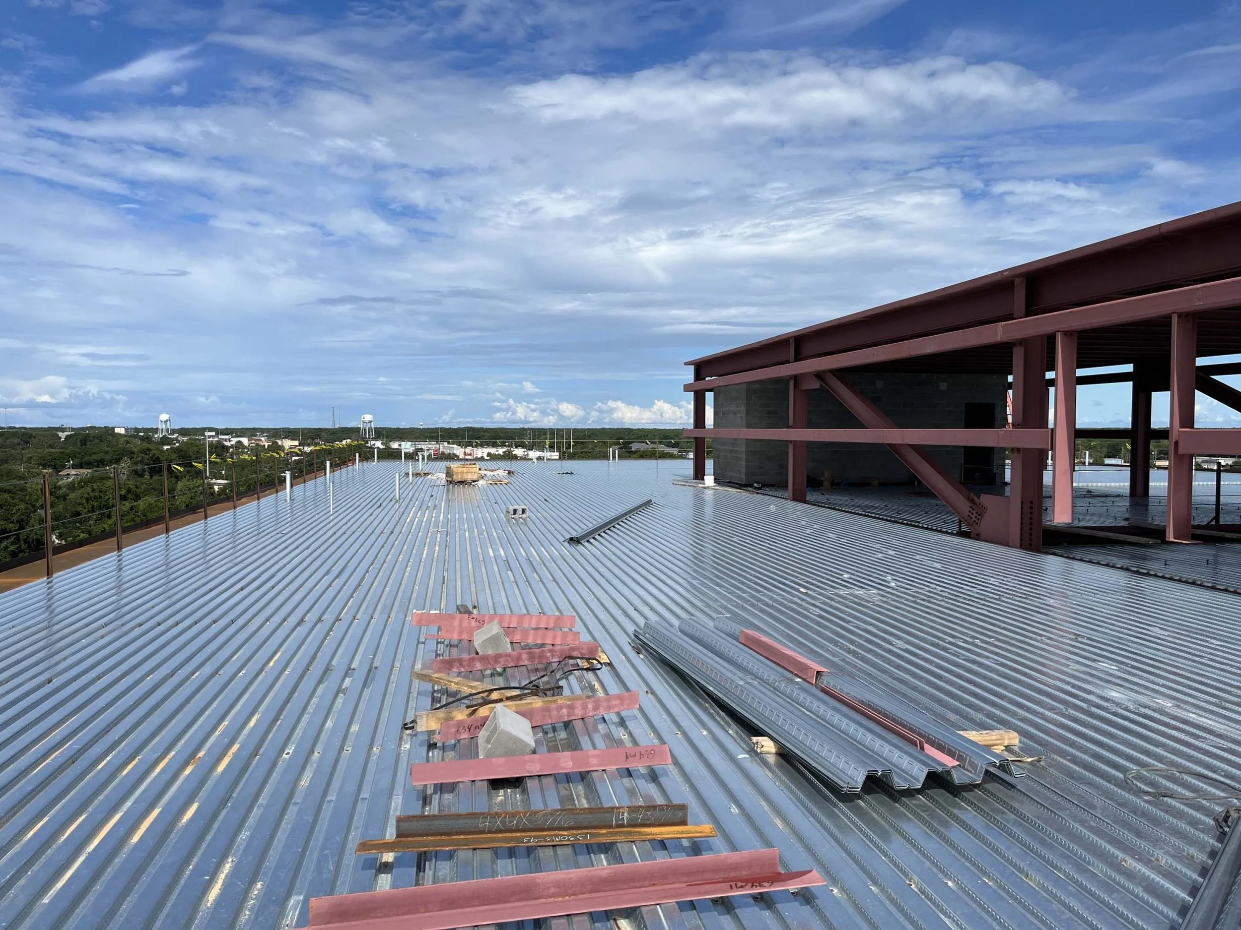 A rooftop under construction with metal panels, building materials, and a partial view of a steel framework structure against a sky with clouds.