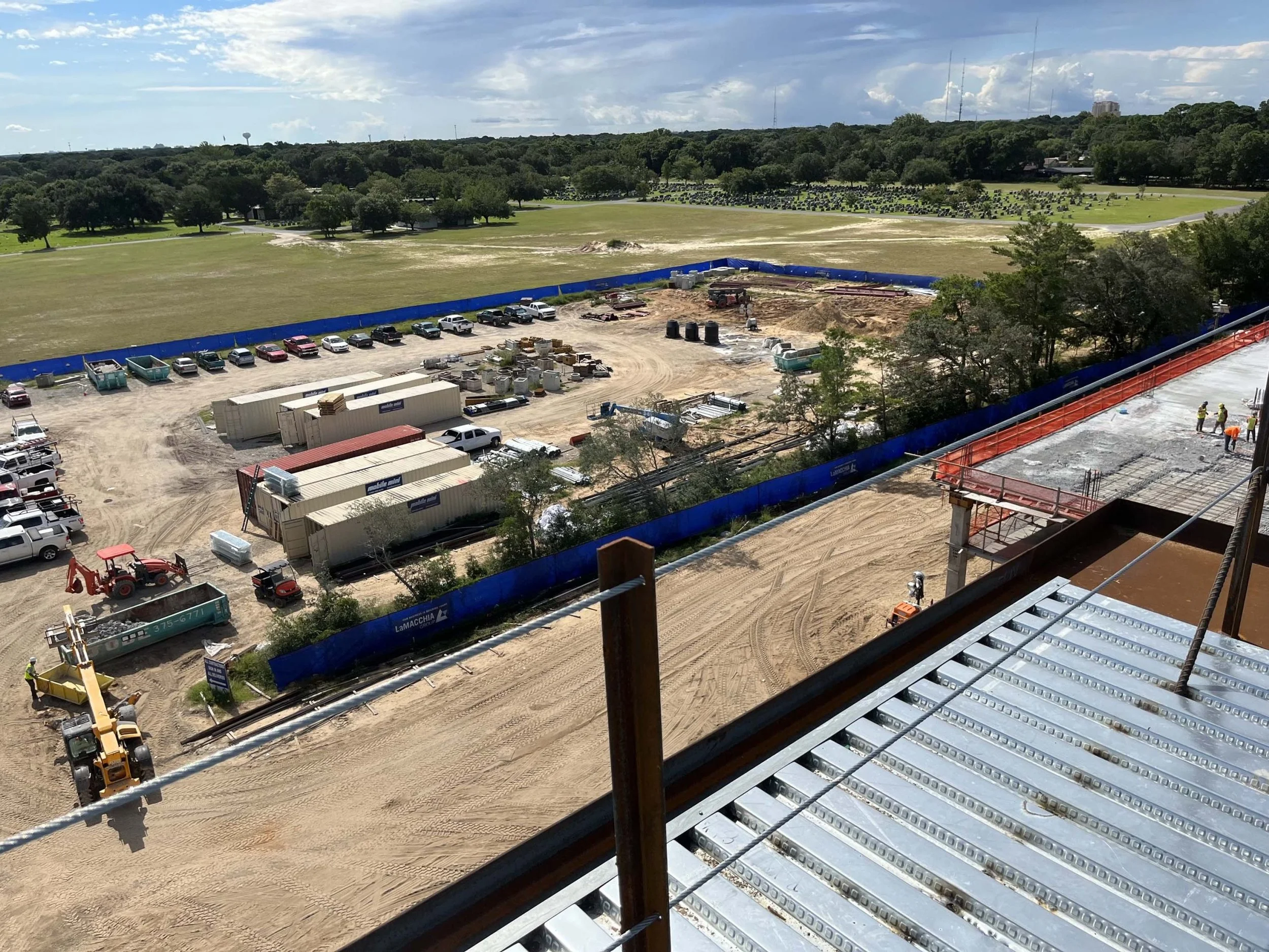 Construction site with workers, construction equipment, and vehicles, enclosed by blue barriers, with a grassy field and trees in the background.