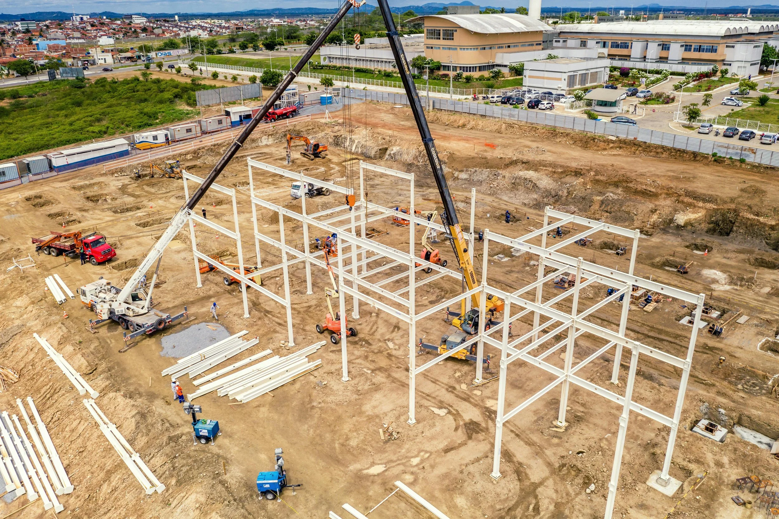 Construction site with steel framework being assembled, cranes, construction vehicles, and workers, surrounded by a fence and nearby buildings.