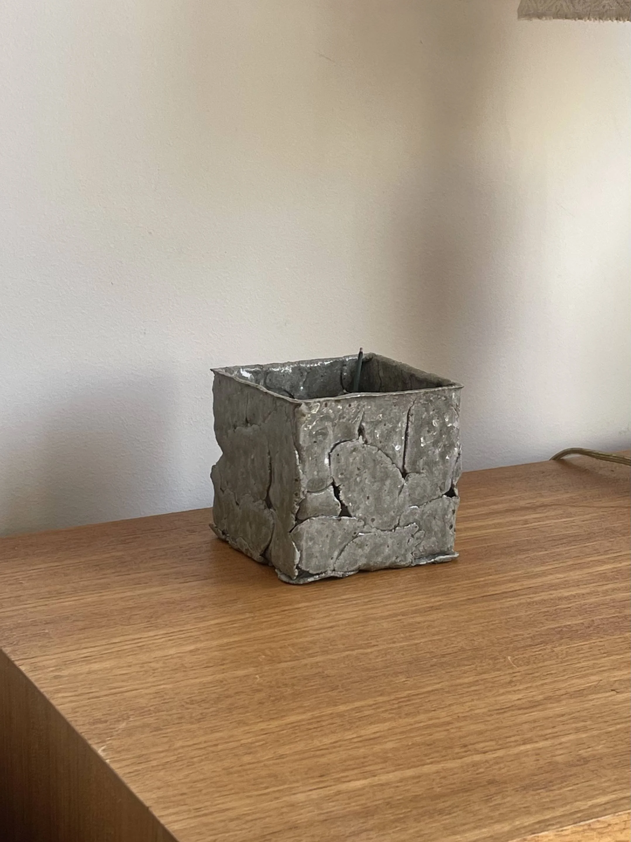 A small, square ceramic incense burner with a rough, cracked surface, sitting on a wooden surface against a plain, off-white wall.