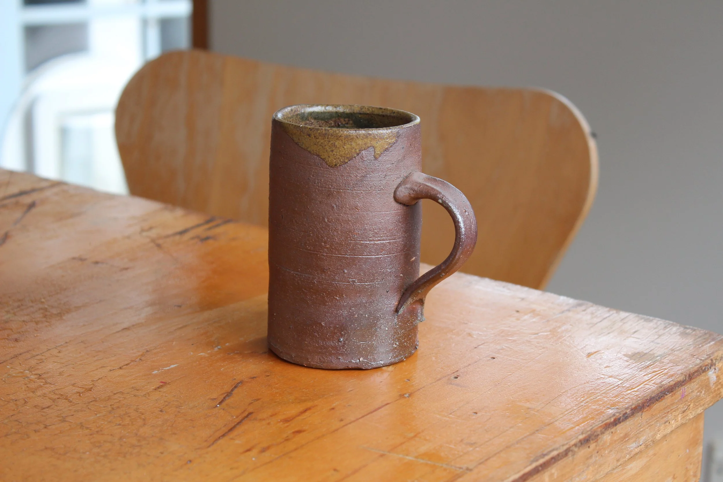 A rustic ceramic mug with a handle sits on a wooden table near a chair. The mug has a textured surface with a natural, earthy color and a slightly uneven glaze at the top.
