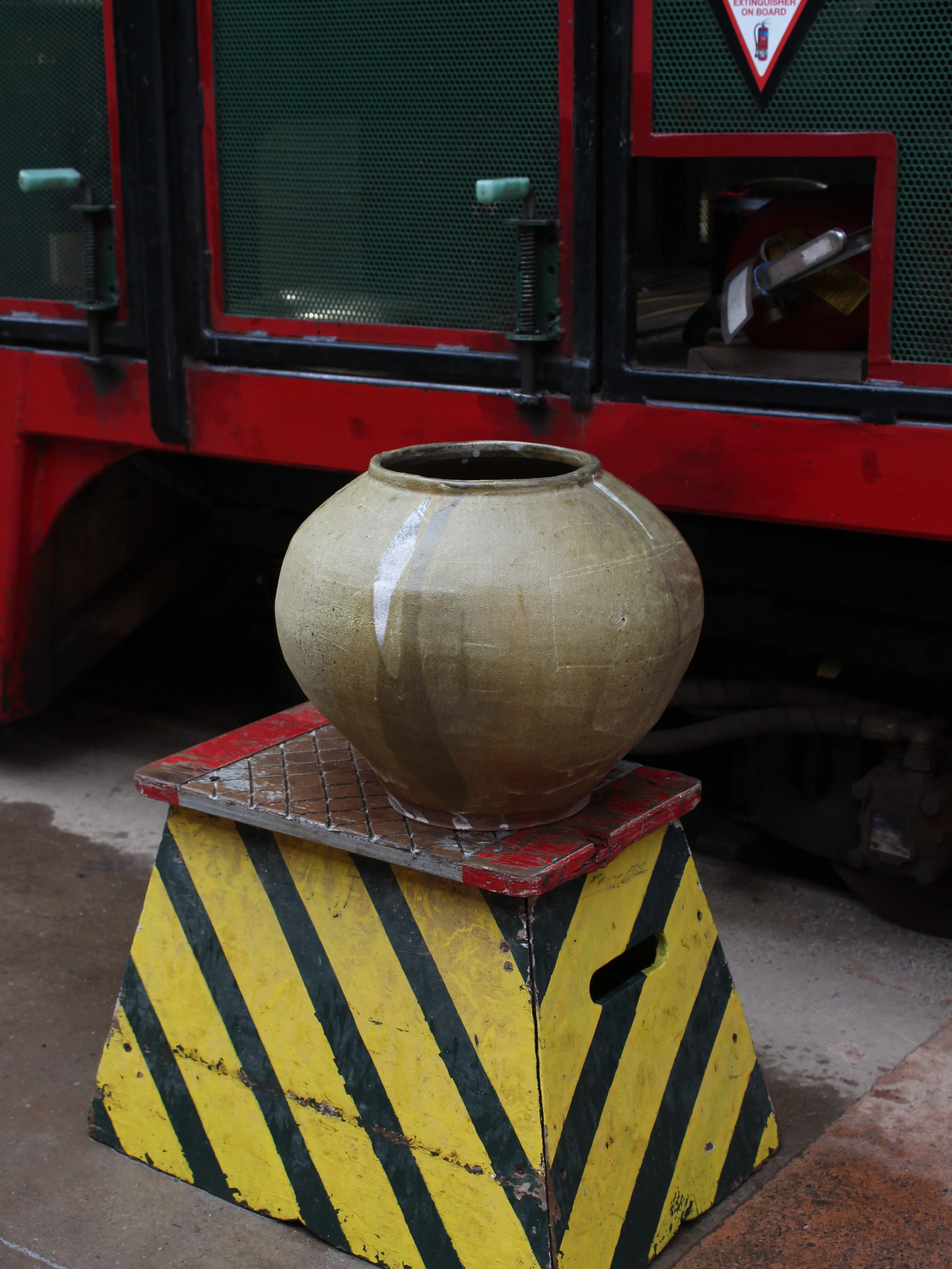 A beige ceramic pot resting on a red and black metal stand with yellow and black striped caution markings, in front of a train.