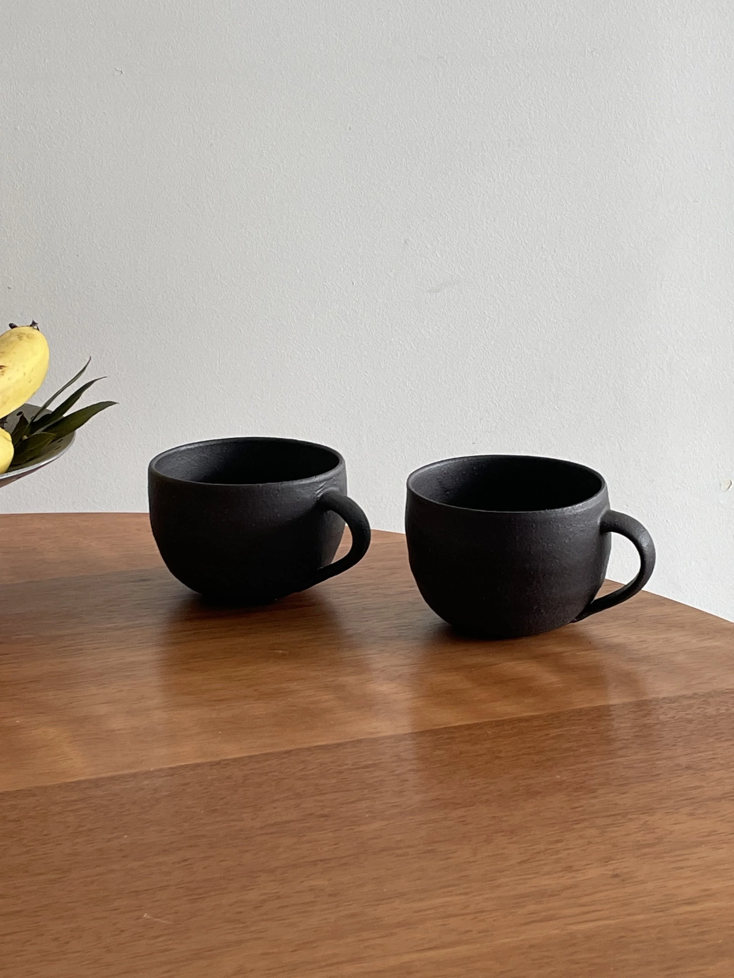 Two black ceramic coffee cups on a wooden table, with a white wall in the background. Part of a bowl with yellow bananas and green leaves is visible on the left side of the image.