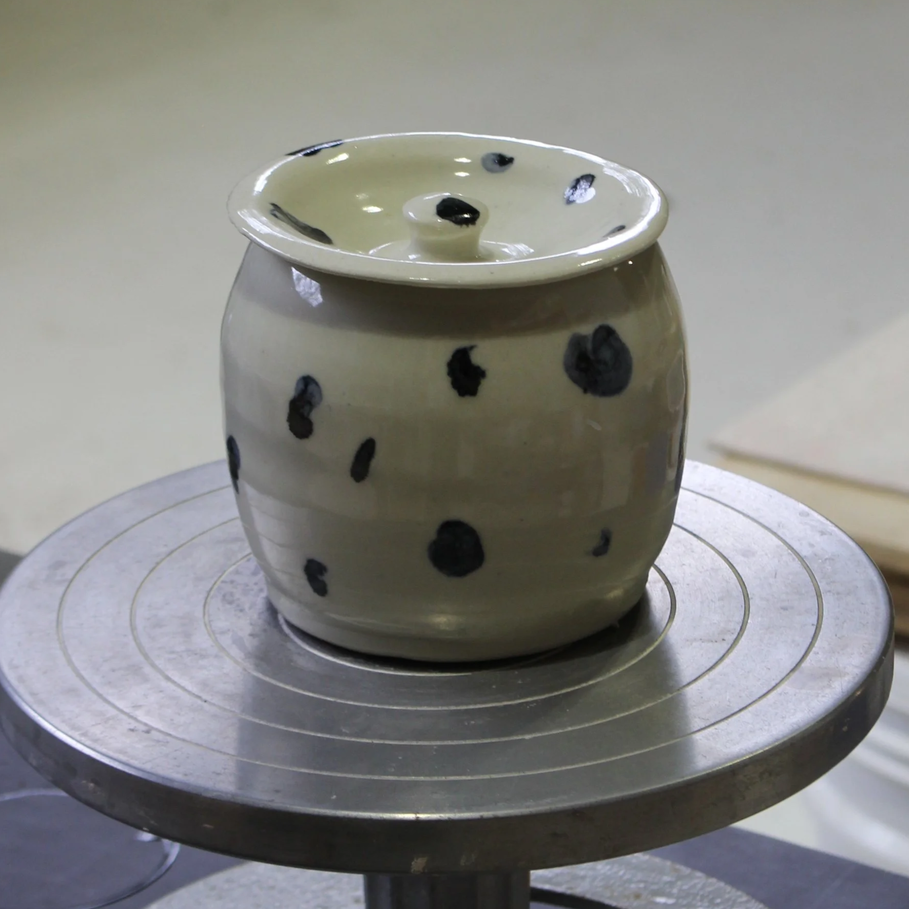 A white ceramic pot with blue spots sits on a rotating wheel in a pottery studio.