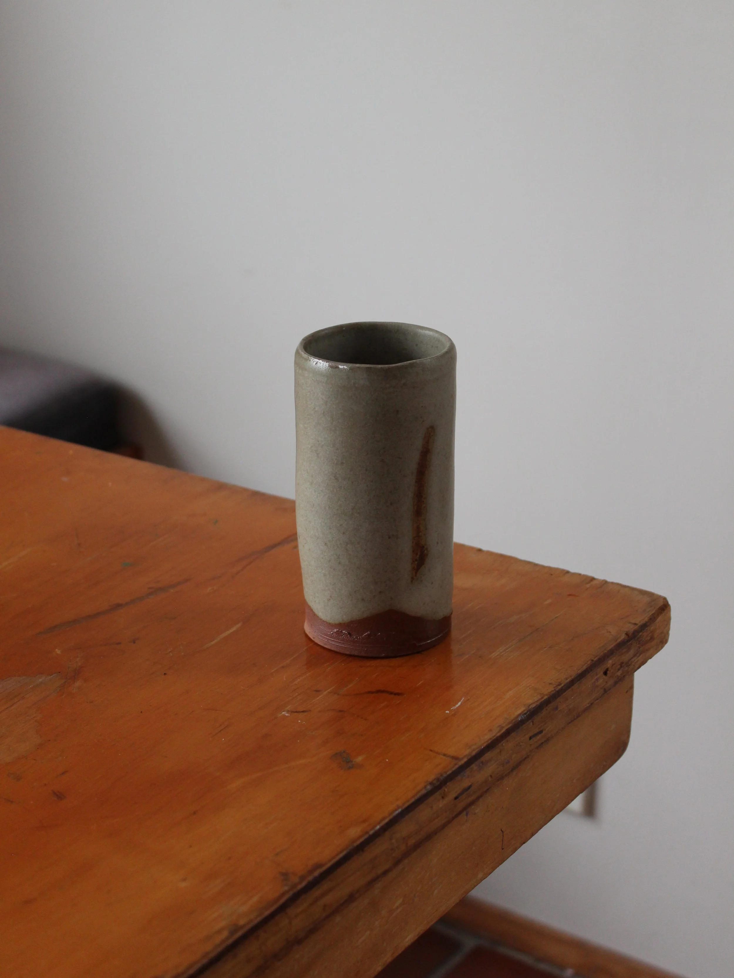 A tall, cylindrical ceramic cup with a beige glaze and a brown bottom, placed on an orange-brown wooden table with a plain white wall in the background.
