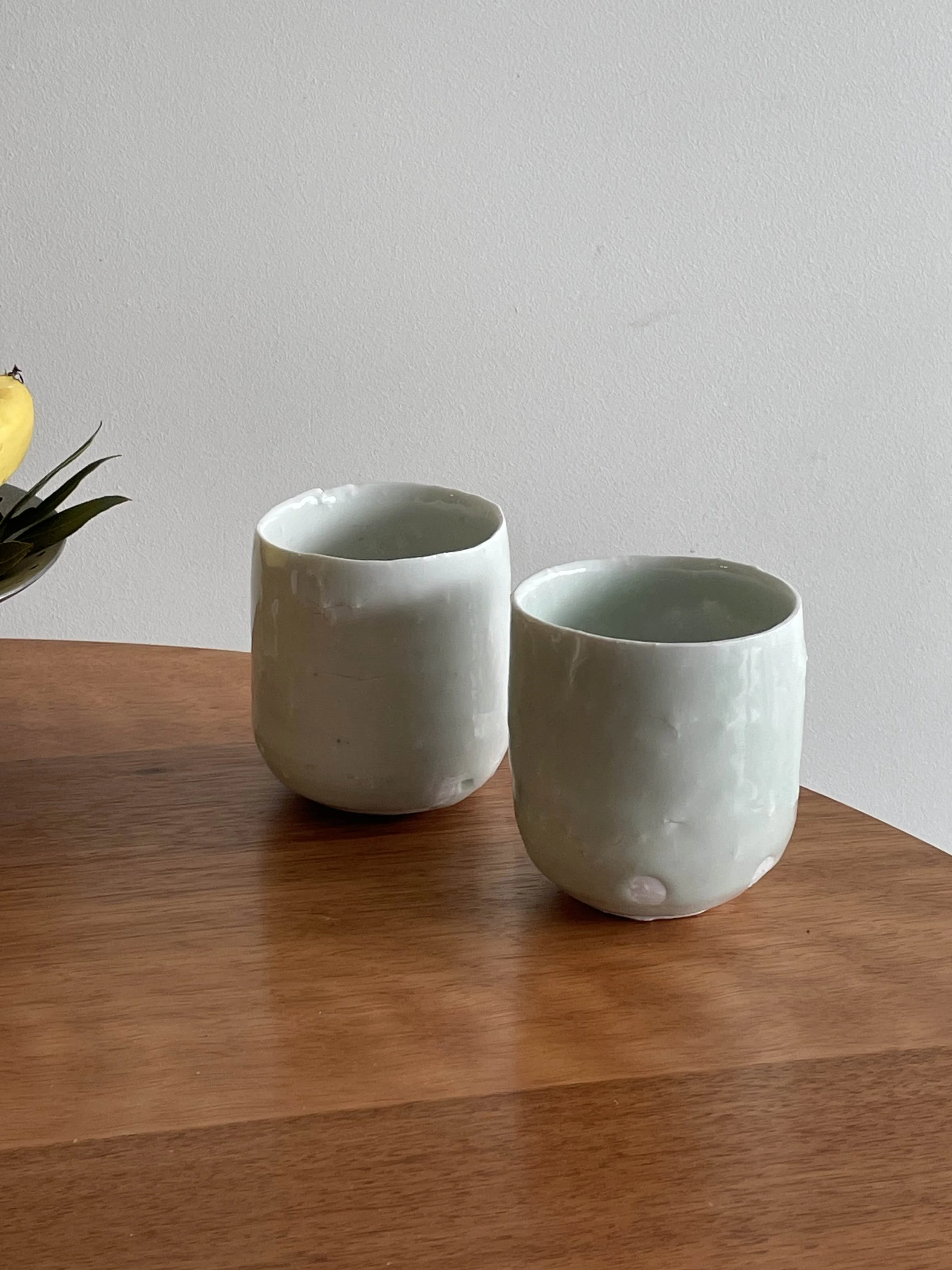 Two white, irregularly shaped ceramic cups on a wooden surface with a plain, light-colored wall in the background.