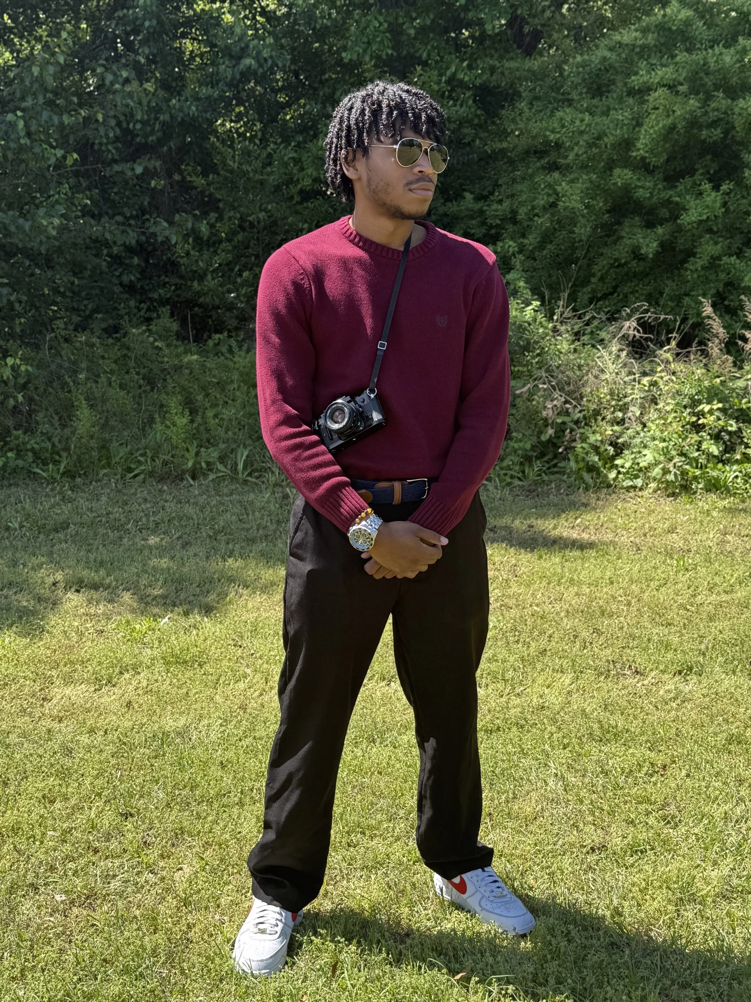 A young man standing outdoors on grass, wearing sunglasses, a maroon sweater, black pants, white sneakers with red accents, a wristwatch, and a camera hanging around his neck. Background includes trees and bushes.