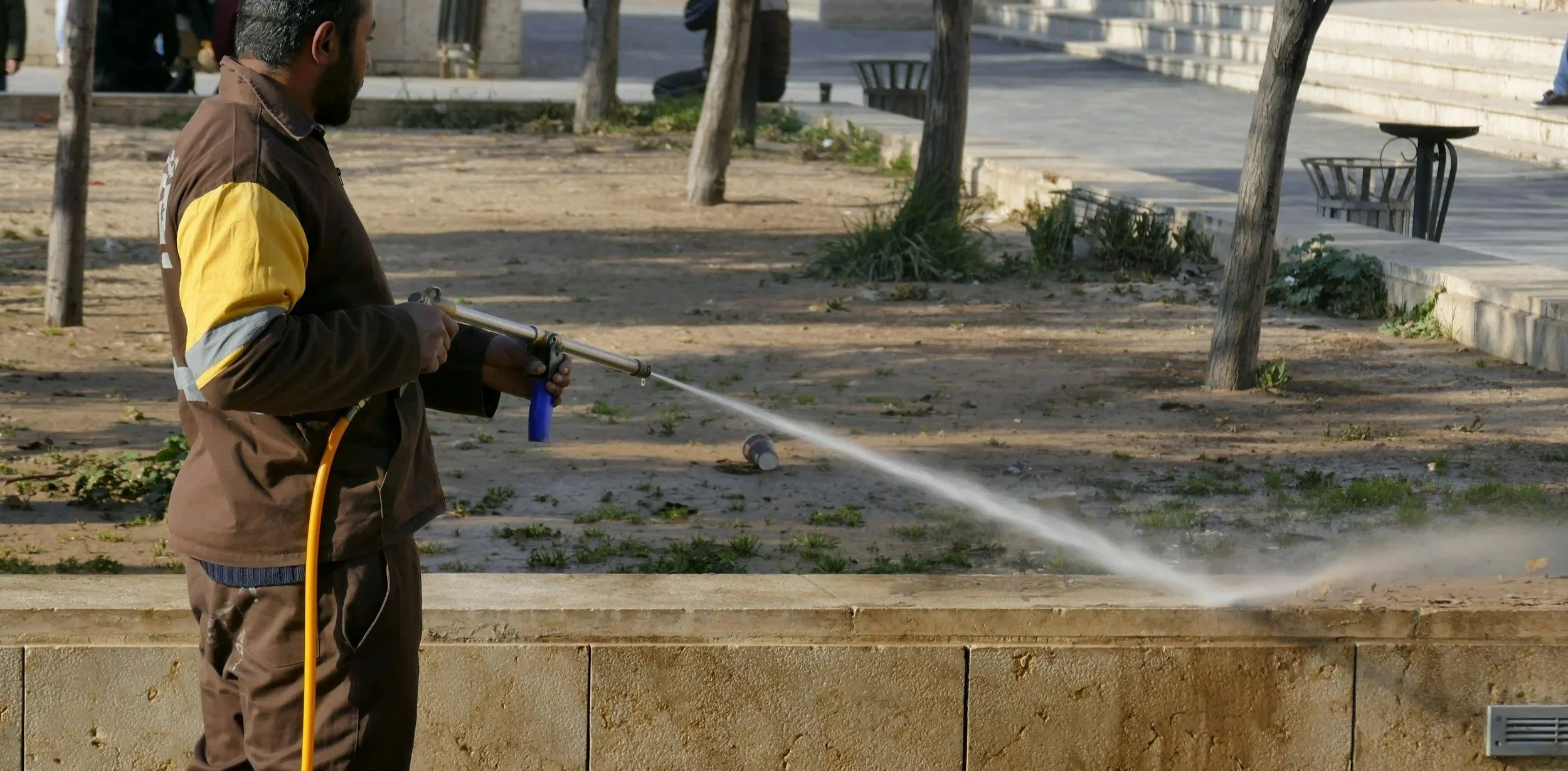 A man is using a pressure washer to clean a concrete surface outdoors, with trees, steps, and benches in the background.
