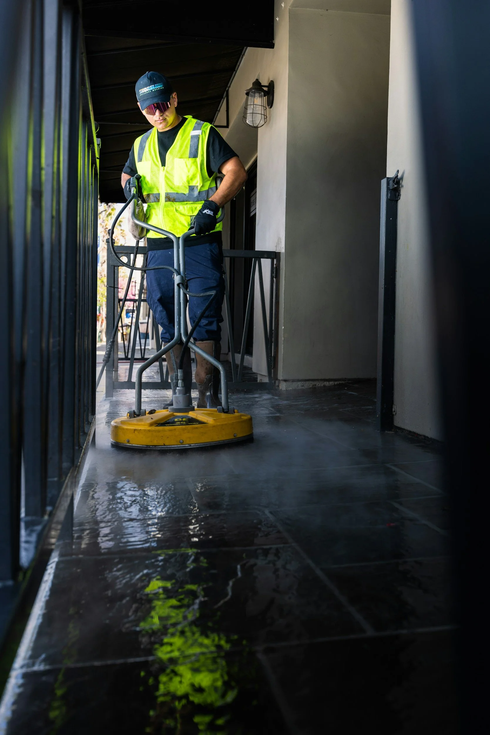 A worker in a yellow safety vest and sunglasses is using a floor buffer to polish outdoor tiles on a balcony.