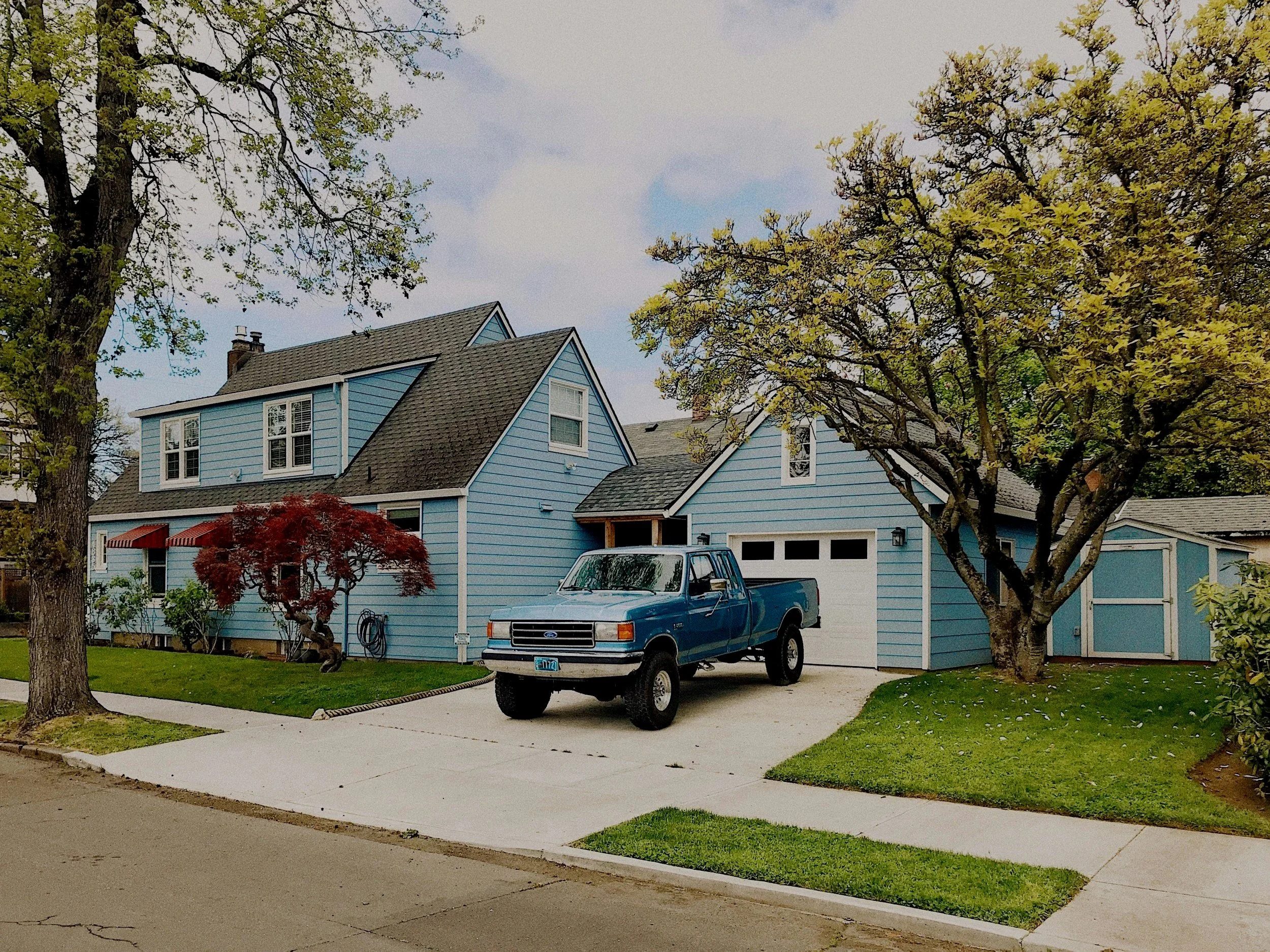 A blue house with a white garage door, surrounded by trees with green and red leaves, and a blue pickup truck parked on the driveway.